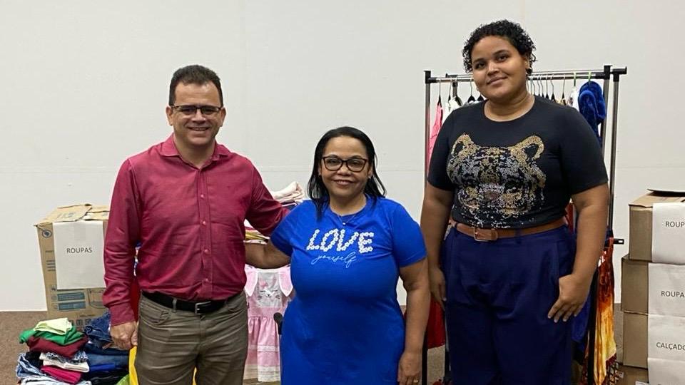 Homem de camisa vinho e calça bege abraça mulher de vestido azul. Ao lado dela, uma moça alta, de camiseta preta e calça azul escura. Eles estão posando para foto de entrega de doação de itens pra crianças e adolescentes