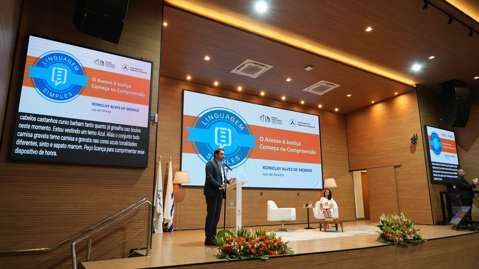 Homem branco, de cabelos castanhos, barba e óculos de grau, fala ao púlpito. Ao fundo, telão projeta imagem sobre linguagem simples. O palco é todo revestido em madeira em tom claro