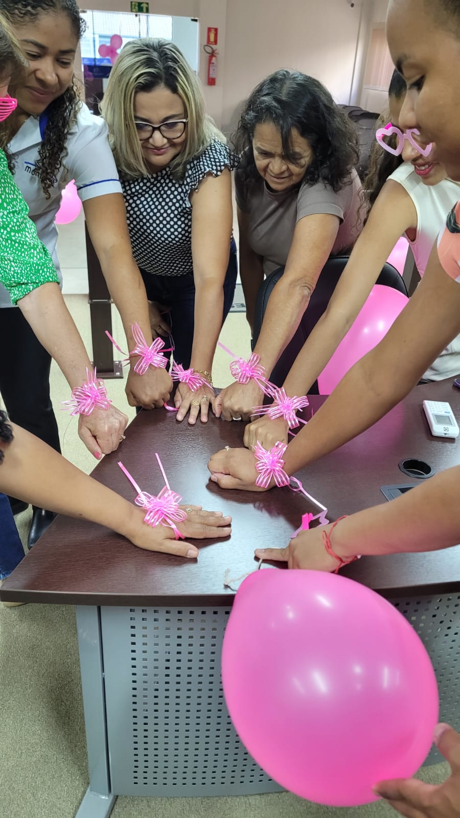 Imagem de um  grupo de mulheres reunido em volta de uma mesa, com as mãos estendidas para o centro, em gesto de união. Todas usam laços cor-de-rosa nos punhos, símbolo da campanha Outubro Rosa, e demonstram alegria e engajamento. Algumas seguram balões também rosas, reforçando o clima de descontração e solidariedade. 