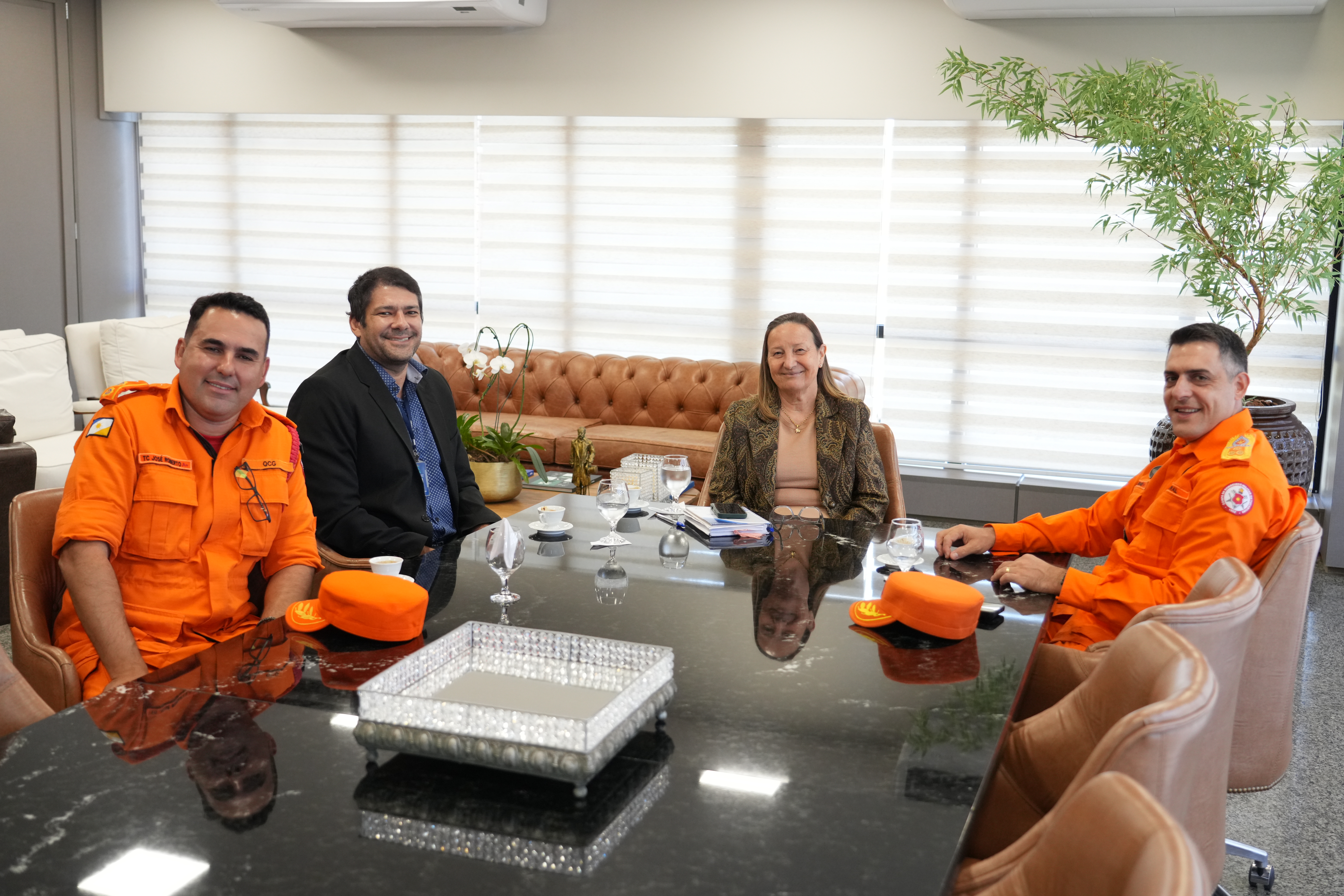 A imagem mostra quatro pessoas sentadas em torno de uma mesa de reunião: dois homens vestem uniformes laranja, do Corpo de Bombeiros, um homem usa blazer preto e camisa azul, e uma mulher veste blazer marrom; sobre a mesa há bonés laranja, copos, documentos e uma bandeja decorativa, com iluminação suave e natural e fundo desfocado que revela um sofá de couro, persianas e uma planta.