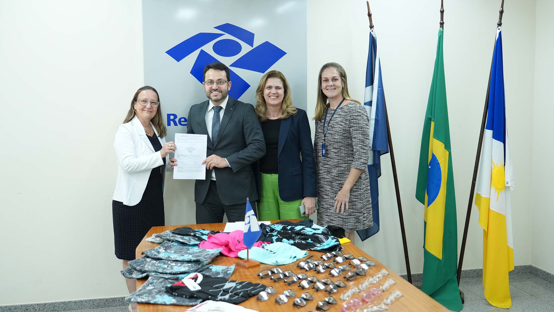 Mulher branca, de vestido preto e blazer branco, cabelos soltos e óculos de grau, segura documento junto com homem branco, de cabelos e barba castanhos, terno e gravata cinza escuro e camisa branca. Ao lado dele, duas mulheres loiras, de cabelos médios, soltos posam juntos para foto. Ao fundo, logo da Receita Federal e mastro com bandeiras em momento de doação de óculos de grau e camisetas posicionados na mesa à frente deles