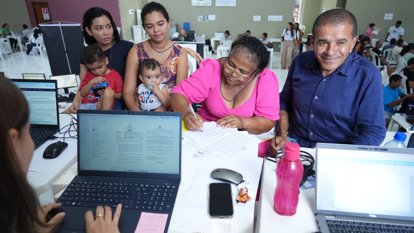 A imagem mostra uma mediação de conciliação de reconhecimento de paternidade pós-morte. No enquadramento, aparece uma pessoa de costas, enquanto quatro adultos e duas crianças estão de frente, e várias outras pessoas ao fundo são atendidas durante a ação.