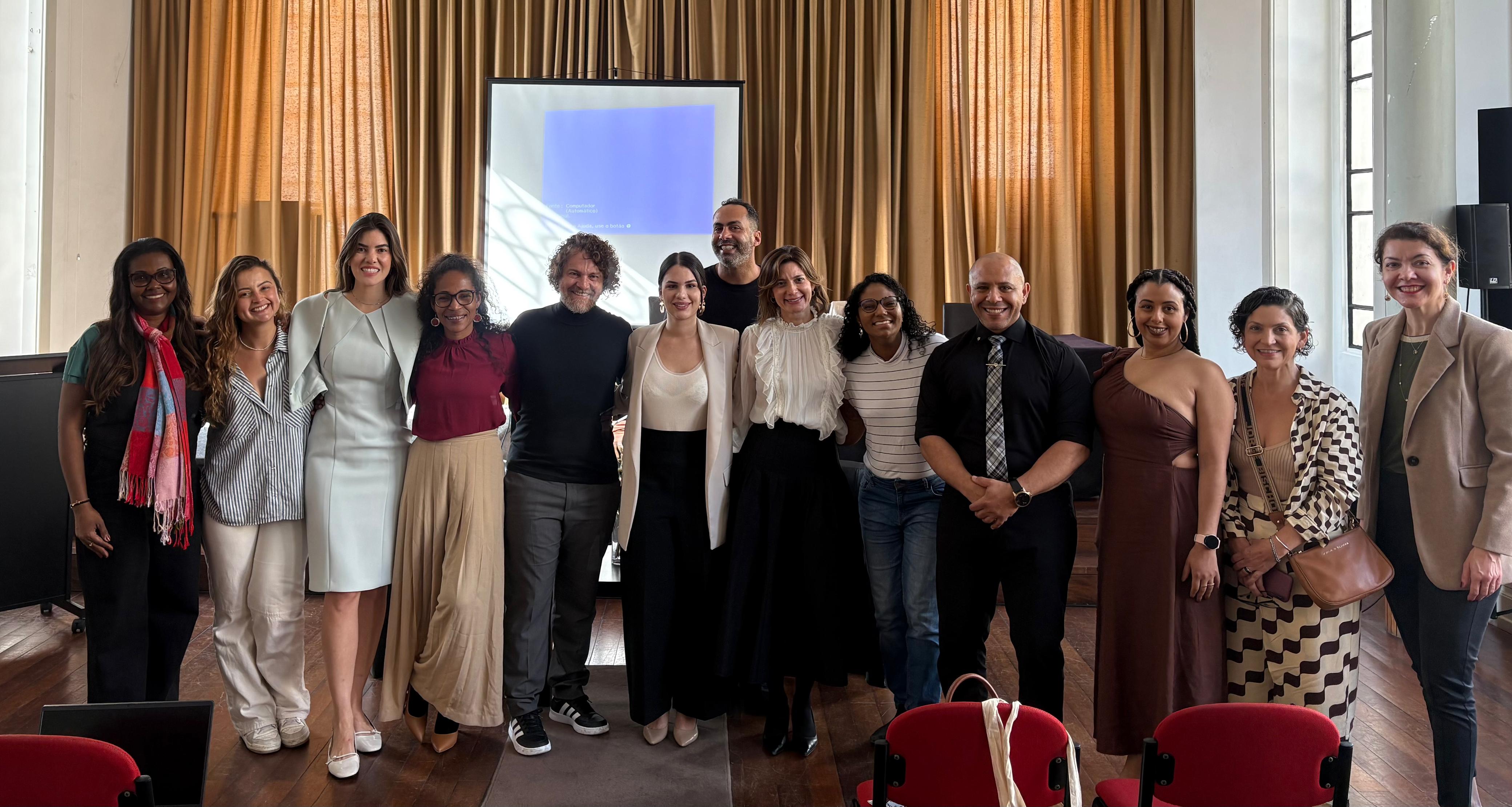Na foto, um grupo de participantes posa sorridente em um ambiente de auditório, com cortinas douradas ao fundo e uma tela de projeção ao centro. São 13 pessoas posicionadas lado a lado, entre homens e mulheres, vestidas em trajes sociais e outras em estilo mais casual.