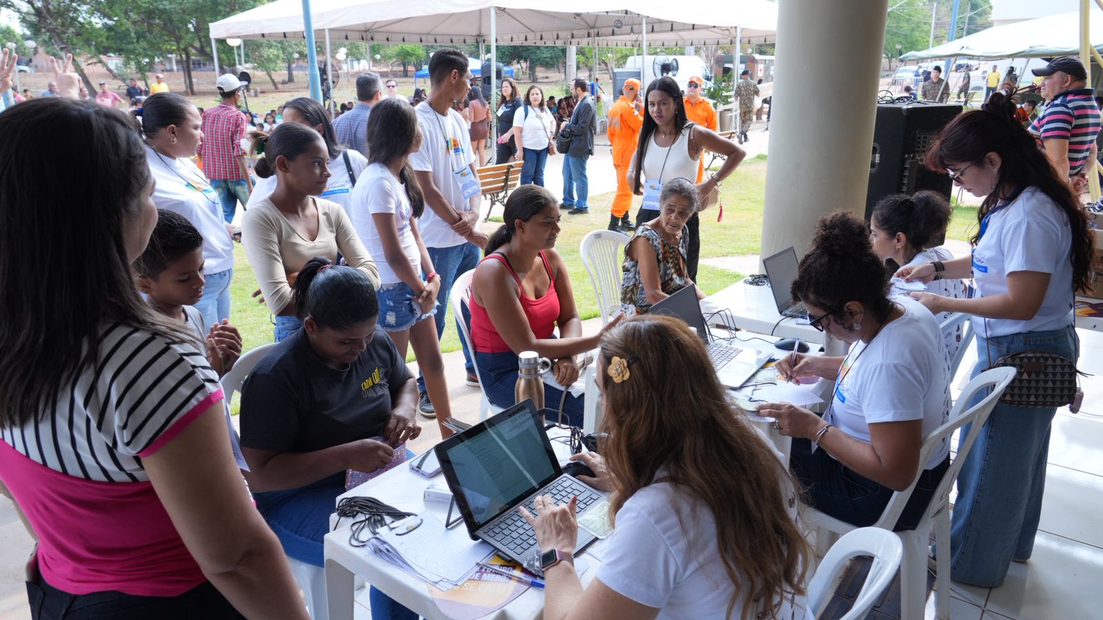 Foto mostra atendentes do Poder Judiciário do Tocantins realizando cadastros e orientações para cidadãos durante uma ação social. As pessoas estão sentadas e em pé, embaixo de uma tenda, aguardando atendimento. No fundo, há outras tendas, servidores e participantes do evento. O ambiente é ao ar livre, com clima de integração e acolhimento.