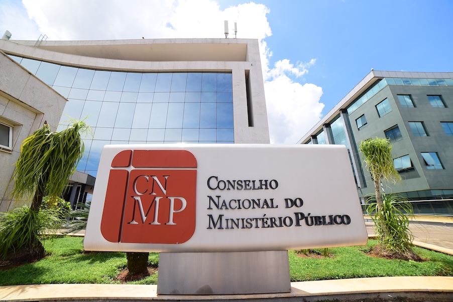 Fachada do prédio do CNMP; a estrutura é composta por paredes brancas e vidraças espelhadas. à frente um jardim com uma placa com o nome Conselho Nacional do Ministério Público em tons claros e vermelho