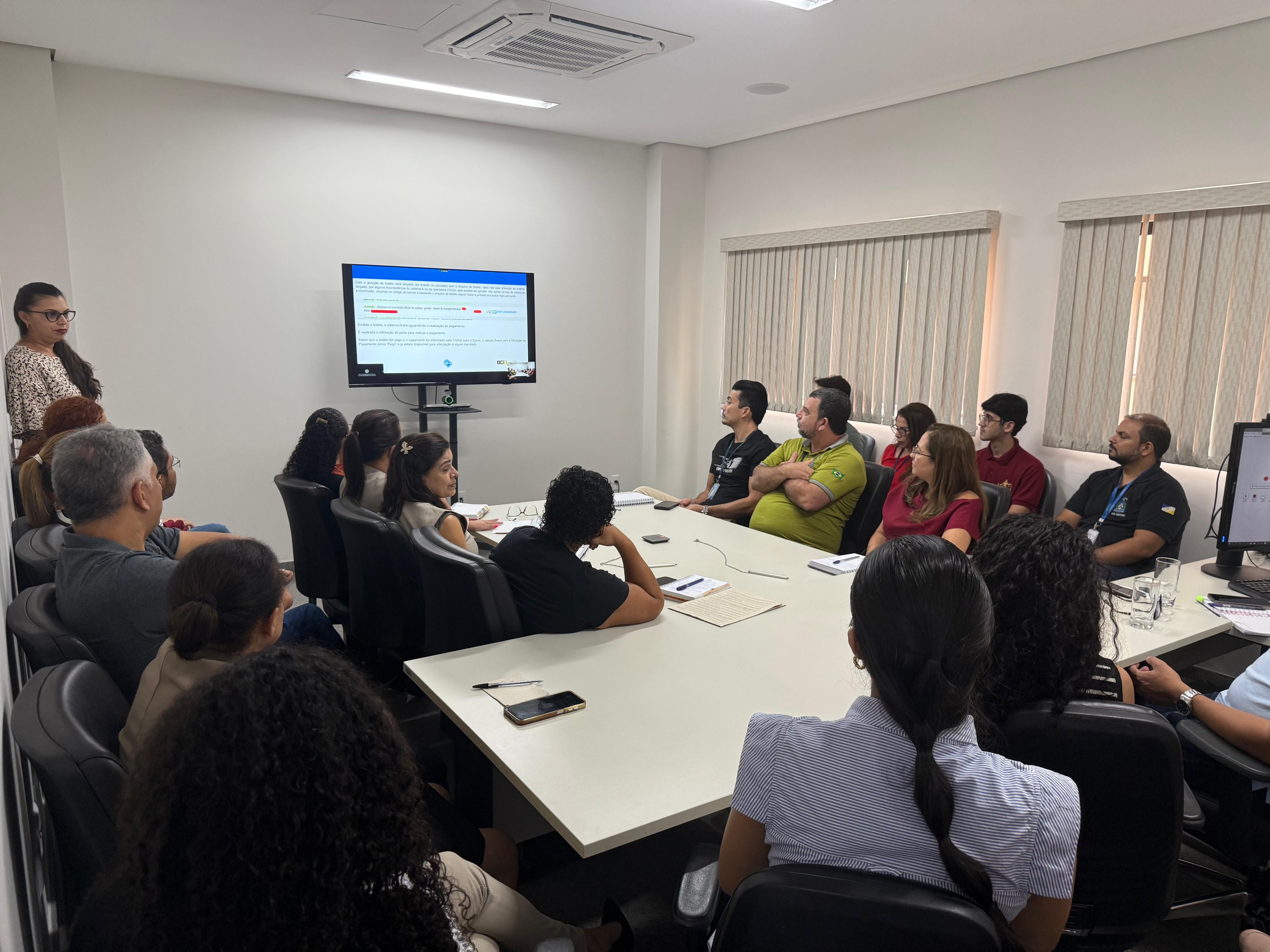 Imagem mostra um grupo de servidores(as) reunidos em uma sala de treinamento do Tribunal de Justiça do Tocantins (TJTO). Eles estão sentados em torno de uma mesa, atentos à apresentação exibida em uma televisão, ao lado da tela tem uma servidora em pé. O encontro tem clima de aprendizado e colaboração, com todos acompanhando as orientações sobre o uso de uma nova ferramenta do sistema eproc. O ambiente é bem iluminado, com janelas e cortinas claras, típico de um espaço de trabalho do Judiciário.
