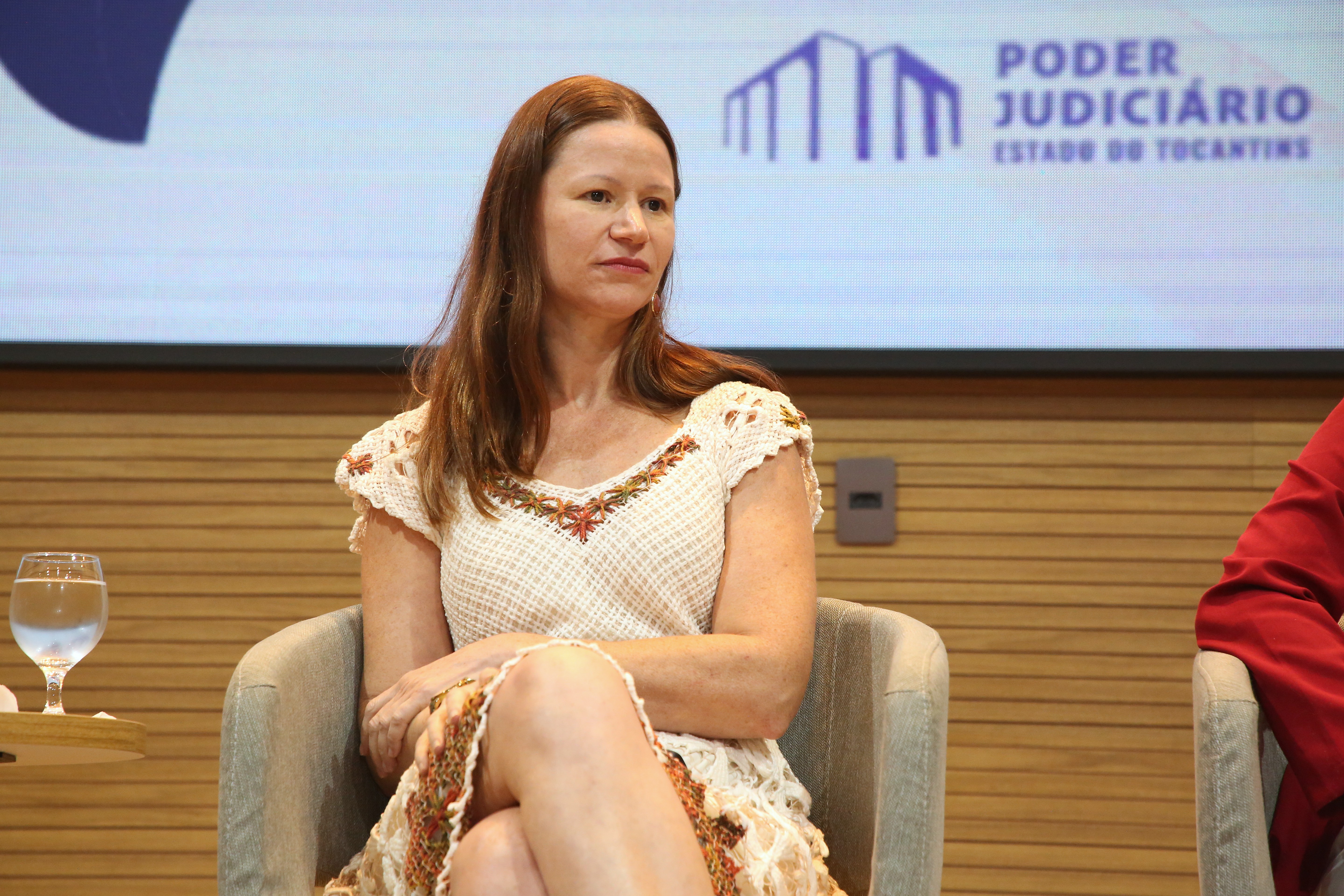 Juliana Biazzi é uma mulher branca, de cabelos claros; ela usavestido claro em tons de laranja e está sentada, no palco do auditório.