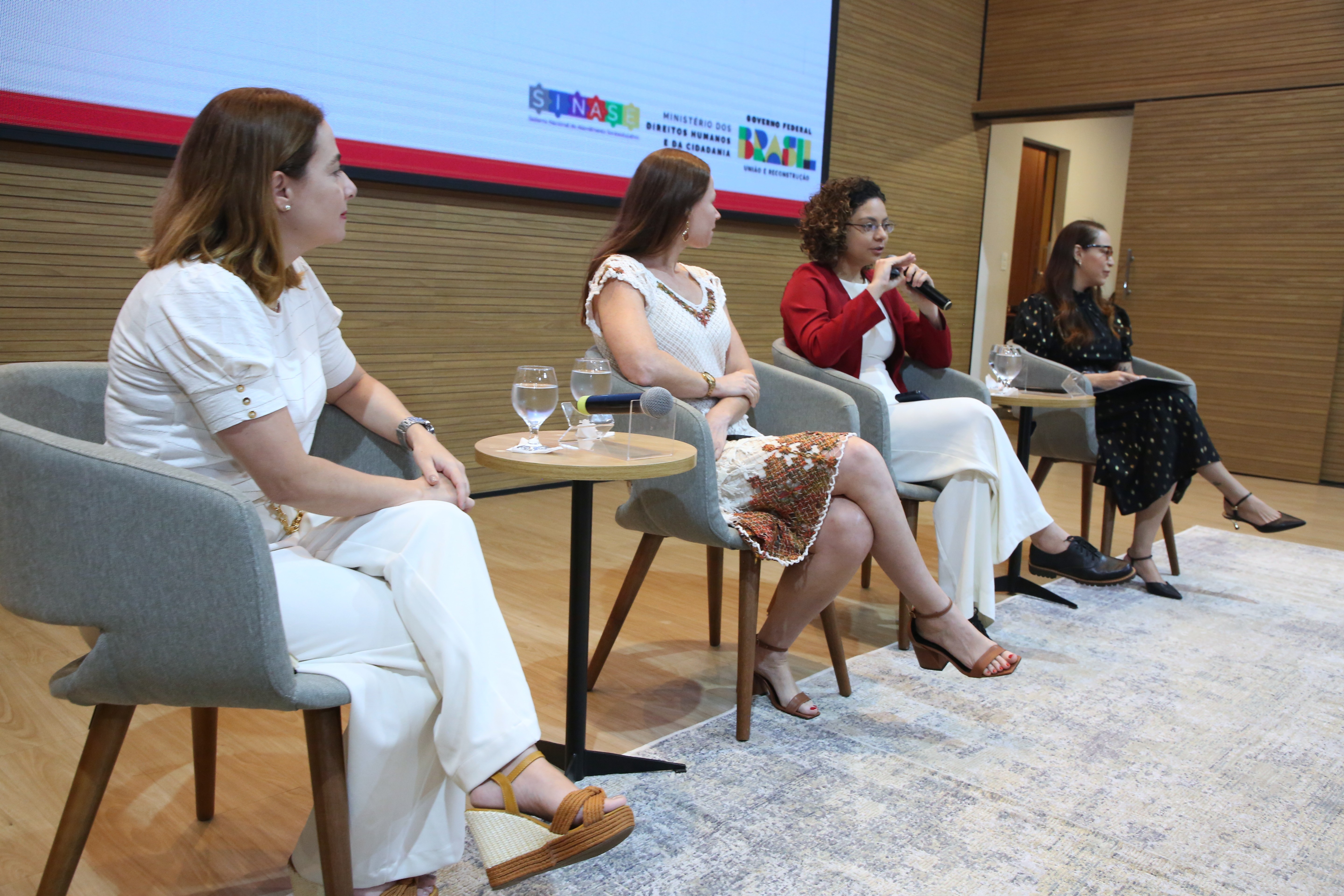 Na imagem, quatro expositoras participam de uma mesa temática durante o evento. Sentadas lado a lado em poltronas cinza, elas dialogam em um ambiente moderno e acolhedor. À esquerda, duas participantes vestem roupas claras, enquanto a terceira, de blazer vermelho e calça branca, fala ao microfone. A quarta expositora, à direita, veste um vestido preto com detalhes dourados e segura um tablet. Ao fundo, o telão exibe os logotipos.