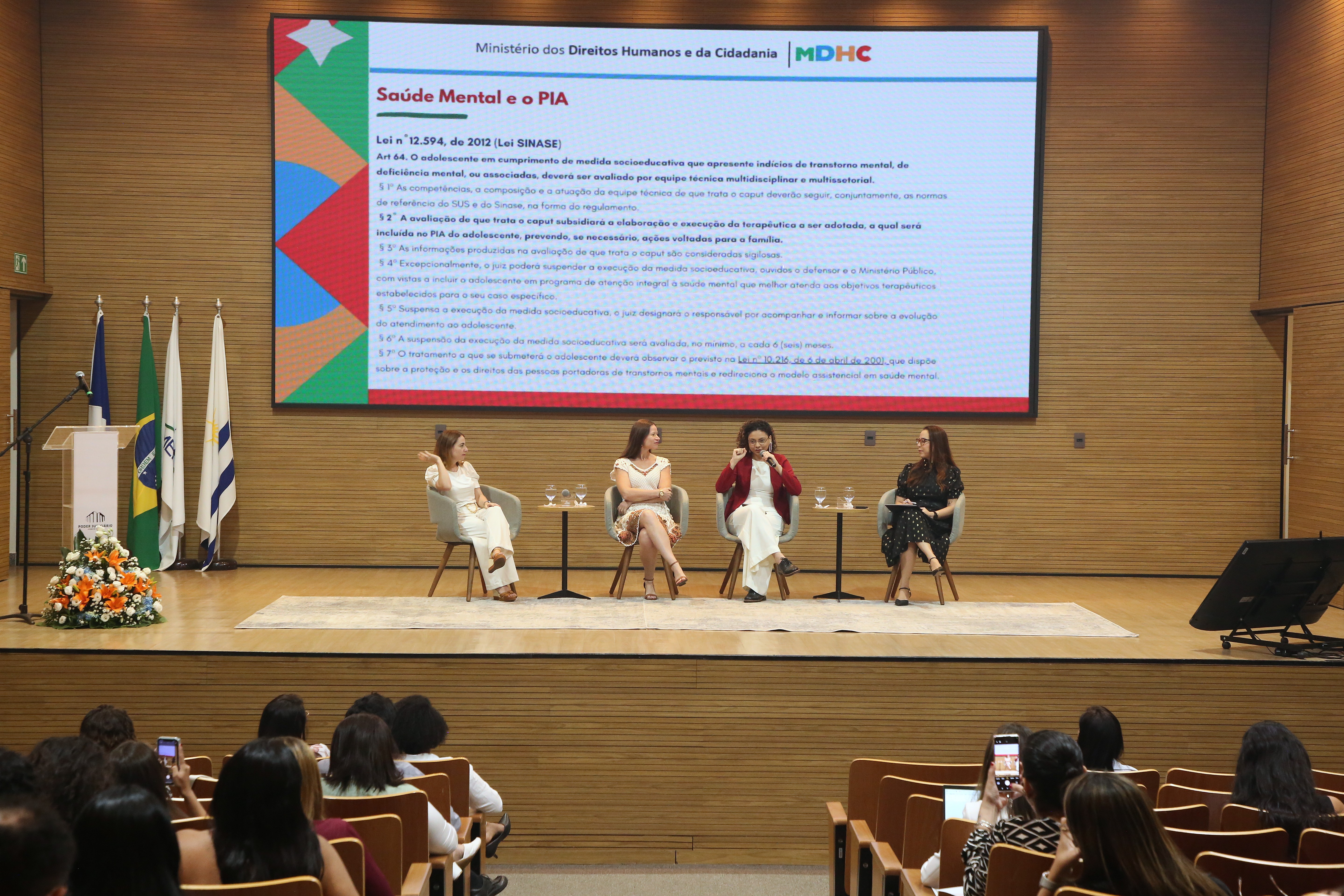 Na foto, quatro expositoras participam de uma mesa temática sobre “Saúde Mental e o PIA”, realizada em um auditório moderno. Sentadas em poltronas cinza no palco, elas dialogam diante de uma plateia atenta. Ao fundo, um grande telão exibe o slide com o título da apresentação e o logotipo do Ministério dos Direitos Humanos e da Cidadania (MDHC), com trechos da Lei nº 12.594/2012 (Lei do Sinase).