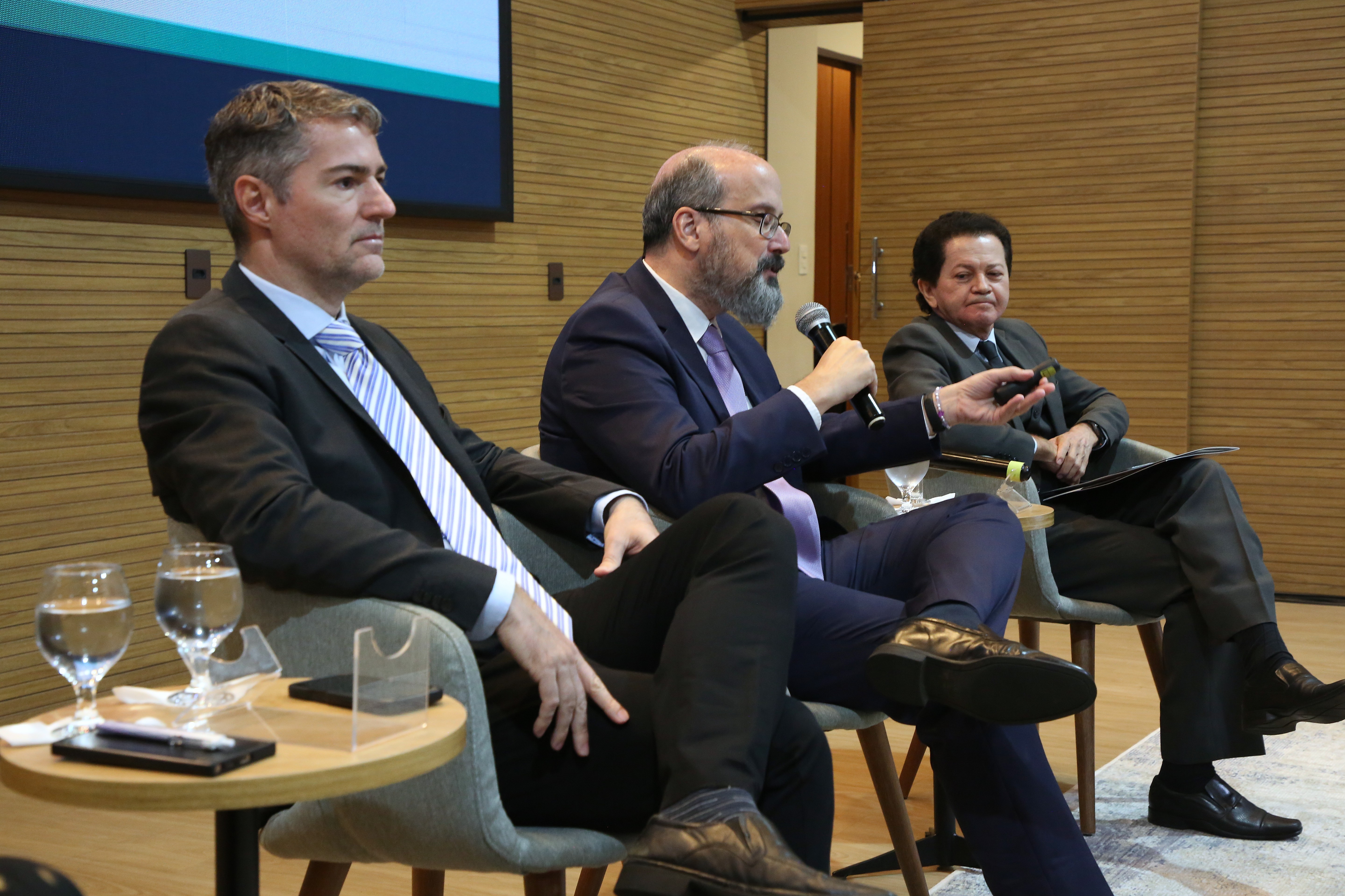 Na foto, três homens participam de uma mesa temática em um auditório. Sentados lado a lado em poltronas cinza, todos vestem terno e gravata, em tom formal e institucional. Ao centro, um dos expositores fala ao microfone, gesticulando enquanto apresenta suas ideias.