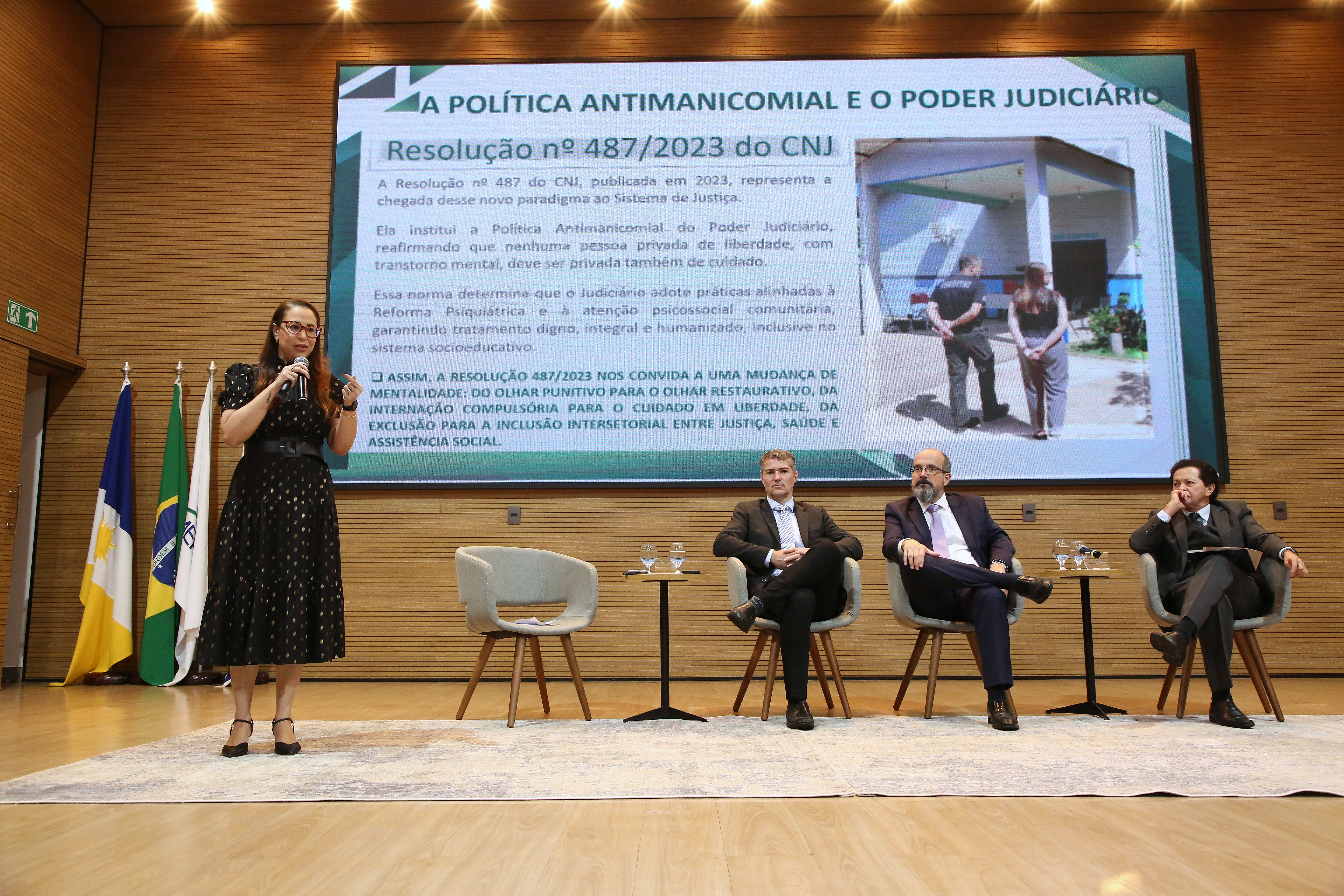 Defensora pública Elisa Maria apresenta sua fala em pé diante do público, enquanto três homens acompanham atentamente sentados ao lado. Ela segura um microfone e gesticula durante a explicação, tendo ao fundo um grande telão com o título “A Política Antimanicomial e o Poder Judiciário” e trechos da Resolução nº 487/2023 do CNJ.