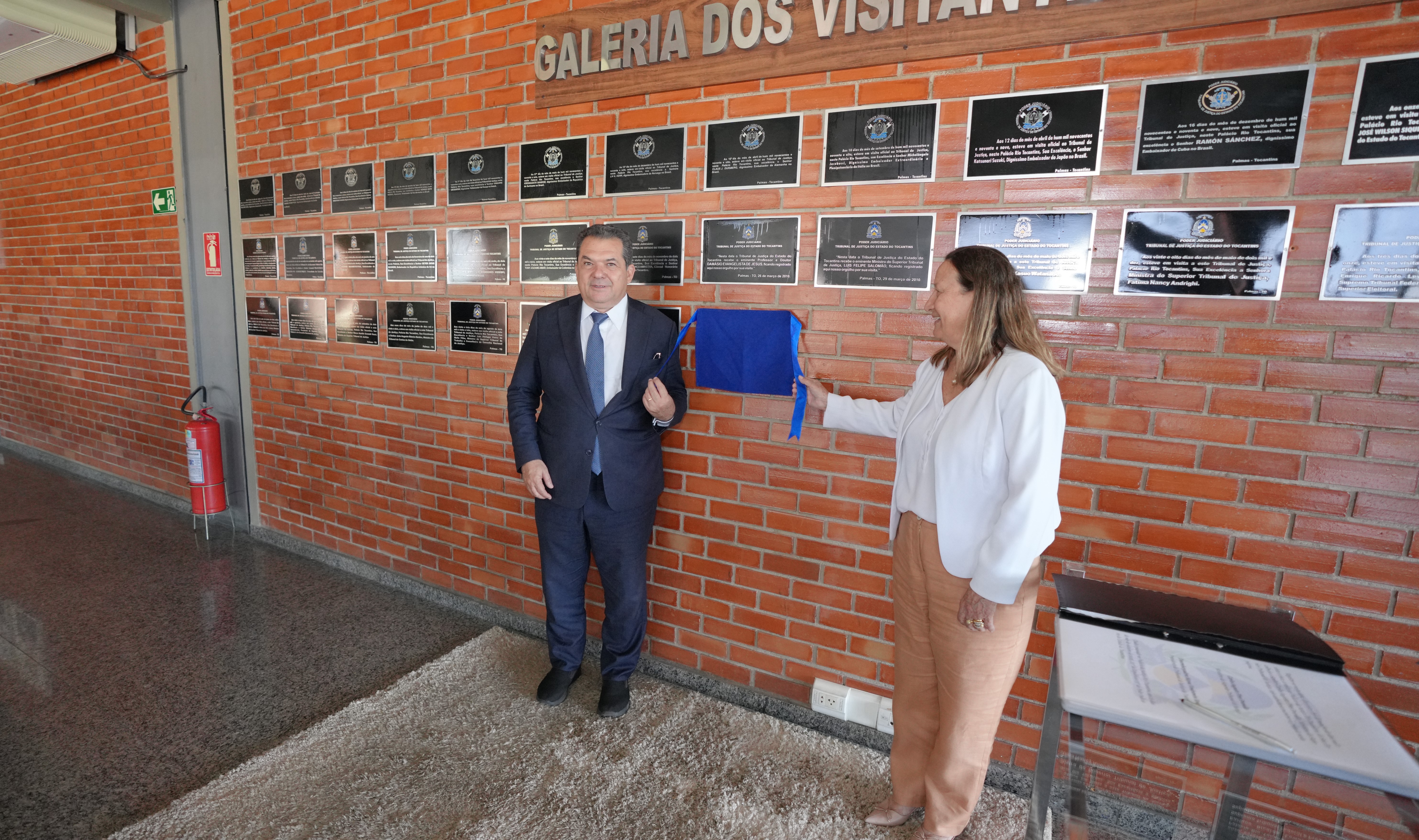 A presidente do Tribunal de Justiça do Tocantins, desembargadora Maysa Vendramini Rosal, e o ministro do Superior Tribunal de Justiça (STJ), Carlos Augusto Pires Brandão, participam do descerramento de uma placa na Galeria dos Visitantes Ilustres do TJTO. Eles estão sorrindo, em frente a uma parede de tijolos com várias placas metálicas, simbolizando o reconhecimento de autoridades que já visitaram o Tribunal.