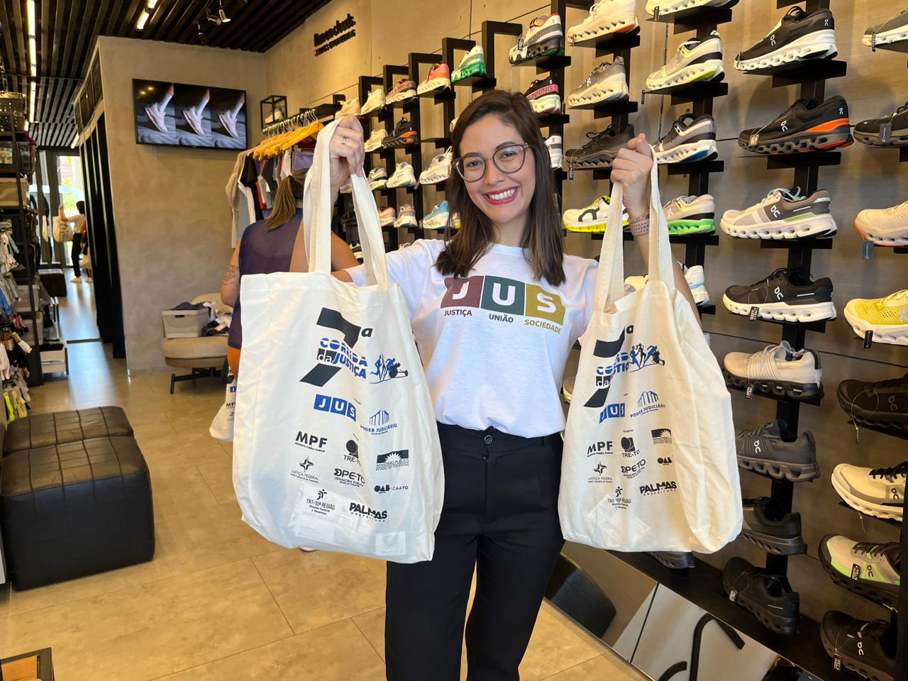 A imagem mostra uma mulher segurando duas sacolas com os itens da corrida da justiça. Ela está vestindo uma camisa com a logo do JUS e está em uma loja de artigos esportivos.