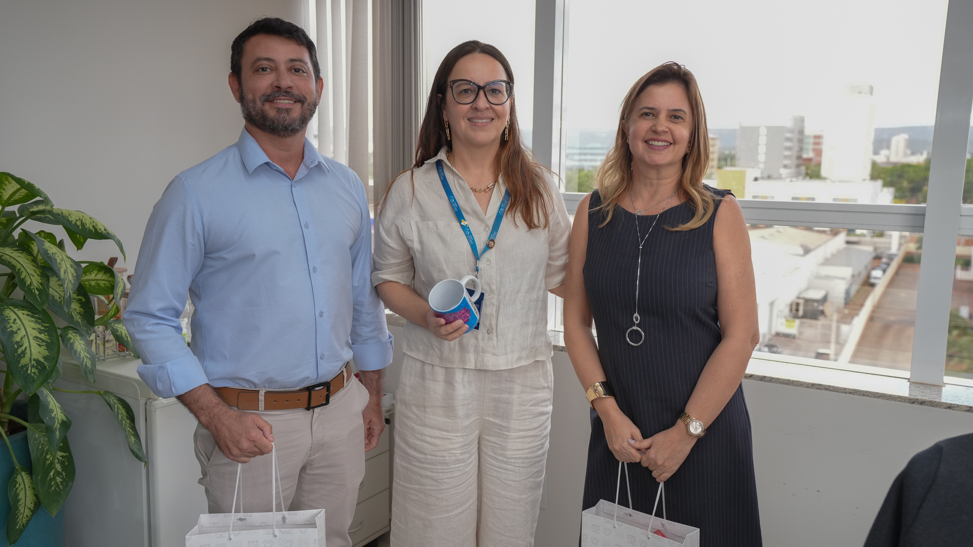 Em uma sala iluminada, três pessoas posam sorridentes para a foto, lado a lado. À esquerda, um homem de barba curta veste camisa azul clara e calça bege, segurando uma sacola branca com a logomarca “JUS”. Ao centro, uma mulher de cabelos castanhos e longos, usando óculos grandes e roupa clara de linho, segura uma caneca colorida e usa crachá azul no pescoço. À direita, uma mulher de cabelos loiros e lisos veste vestido preto sem mangas, com colar e relógio dourados, também segurando uma sacola semelhante. Atrás deles, uma grande janela mostra parte da cidade e um céu claro; à esquerda, há uma planta verde em um vaso azul.