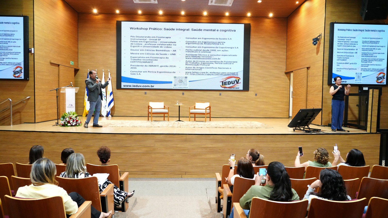 Auditório amplo e moderno, com paredes revestidas de madeira clara e iluminação suave no teto. No palco, um homem de terno cinza fala ao microfone, gesticulando com a mão direita em direção ao telão, onde há uma grande apresentação projetada com o título “Workshop Prático: Saúde Integral – Saúde mental e cognitiva”. À direita do palco, uma intérprete de Libras veste blusa preta e calça azul, traduzindo a fala do palestrante. No centro do palco, há duas poltronas brancas com estrutura de madeira e uma mesinha com copos d’água entre elas. Na plateia, pessoas sentadas assistem e algumas registram o momento com seus celulares. O ambiente transmite organização, acolhimento e atenção ao tema da saúde e bem-estar.