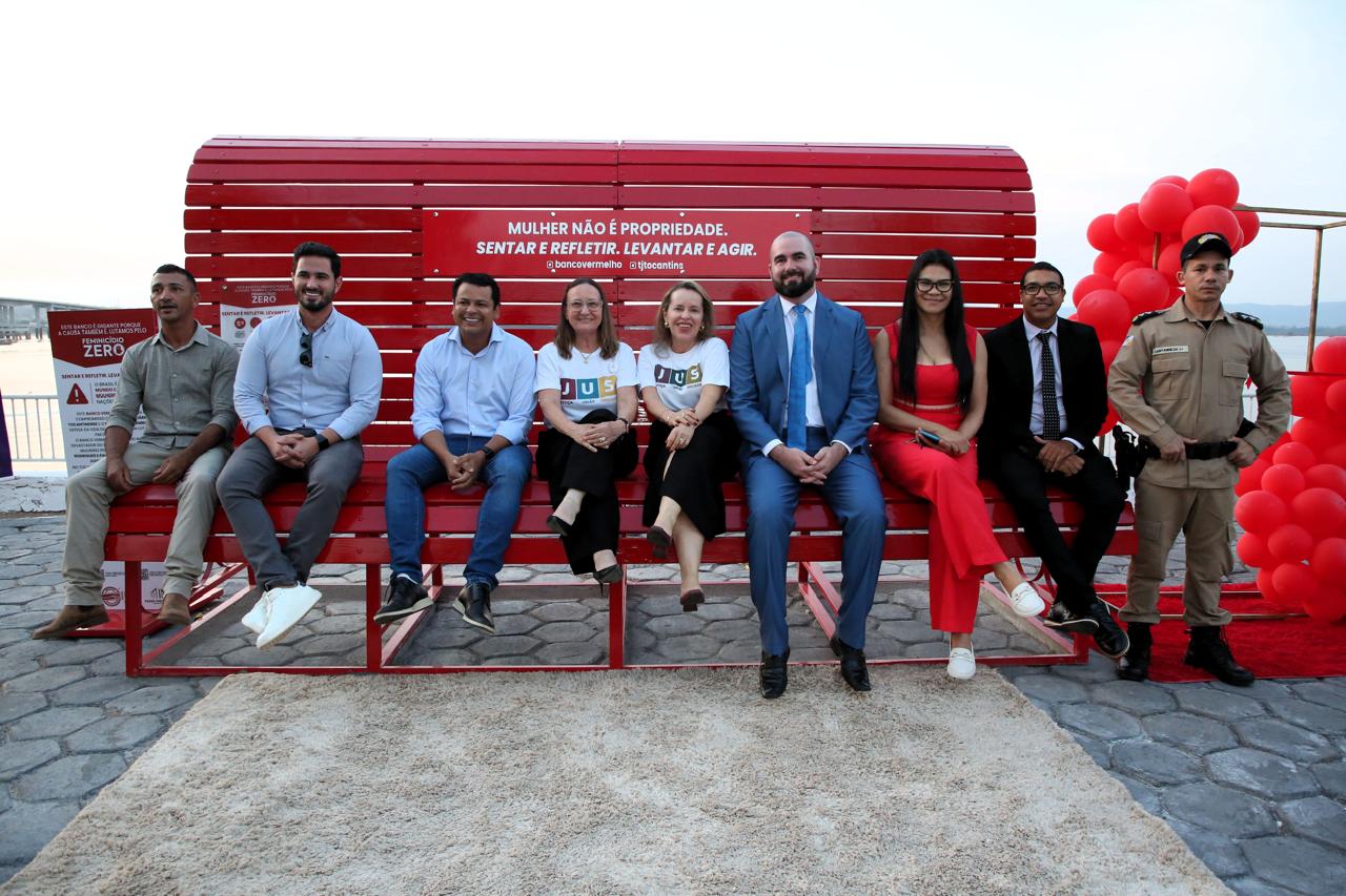 Oito pessoas estão sentadas lado a lado em um grande banco vermelho, instalado em ambiente externo, sobre piso de pedras. O banco exibe a frase em branco: “Mulher não é propriedade. Sentar e refletir. Levantar e agir.” À direita, há um conjunto de balões vermelhos e um homem vestindo um uniforme bege da polícia militar. O céu está claro ao fundo, com uma leve névoa.  O grupo é formado por homens e mulheres de diferentes idades; alguns usam roupas formais, outros, mais casuais. Duas mulheres no centro vestem camisetas brancas do projeto com a palavra “JUS” estampada. Ao lado direito, um homem de terno azul e gravata senta próximo a uma mulher de macacão vermelho vibrante. 