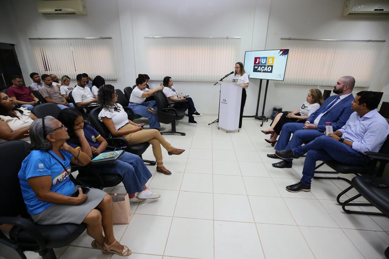 Em uma sala de reuniões iluminada, com paredes brancas e janelas cobertas por persianas, cerca de 20 pessoas estão sentadas em cadeiras pretas, voltadas para a frente. No canto direito,  a presidente do tribunal de justiça, desembargadora Maysa Vendramini Rosal fala ao microfone atrás de um púlpito branco. Ao lado dela, na tela de um monitor, aparece o logotipo colorido do projeto JUS. Na primeira fileira, à direita, três pessoas — duas mulheres e um homem — escutam atentamente; a mulher usa camiseta branca do projeto, o homem está de terno azul. Do lado esquerdo, entre o público, estão homens e mulheres de diferentes idades, alguns de camiseta branca do evento, outros com roupas casuais, acompanhando a fala.