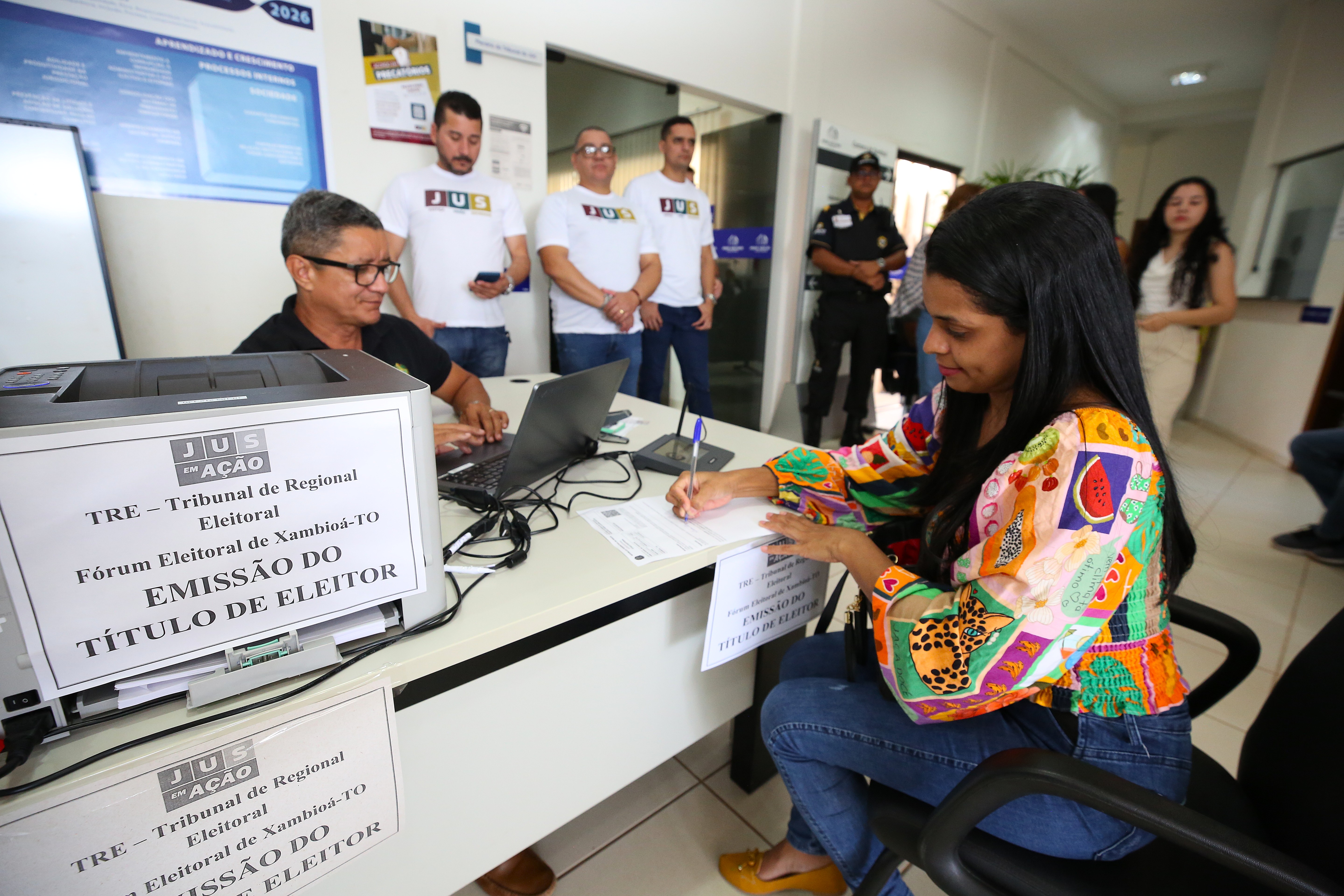 Em um ambiente de atendimento, uma mulher jovem de cabelos longos e lisos está sentada à direita, preenchendo um formulário com caneta sobre a mesa. Ela veste blusa colorida estampada com flores e figuras geométricas e calça jeans. À esquerda, um homem de óculos, camisa preta, opera um notebook atrás da mesa; sobre a mesa e à frente dele há uma impressora com um cartaz que diz “Emissão do Título de Eleitor – TRE / Fórum Eleitoral de Xambioá-TO”. Ao fundo, quatro homens de camiseta branca com o logotipo “Jus em Ação” observam a cena; um deles segura um celular. Há também uma mulher de vestido claro e um policial fardado. O local tem piso claro, paredes brancas e luz natural entrando pelo corredor.