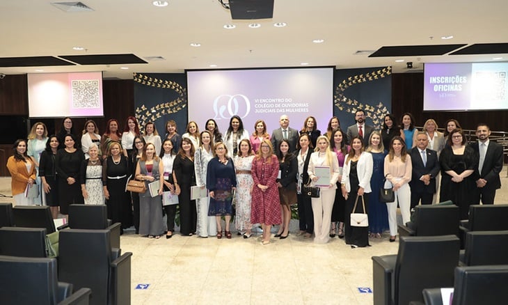 Em um auditório iluminado, um grupo de cerca de 40 pessoas — a maioria mulheres — posa para uma foto oficial em frente a uma tela de projeção. No centro do telão, lê-se: “VI Encontro do Colégio de Ouvidorias Judiciais das Mulheres (Cojum)”.  As participantes estão dispostas em duas fileiras, sorrindo, com trajes formais e elegantes em tons claros, escuros e florais. O ambiente transmite seriedade e acolhimento, com cadeiras dispostas ao redor e painéis decorativos ao fundo. À direita, há um painel com a frase “Inscrições Oficinas”. A imagem simboliza a união e o compromisso das ouvidorias do Judiciário na defesa dos direitos das mulheres.
