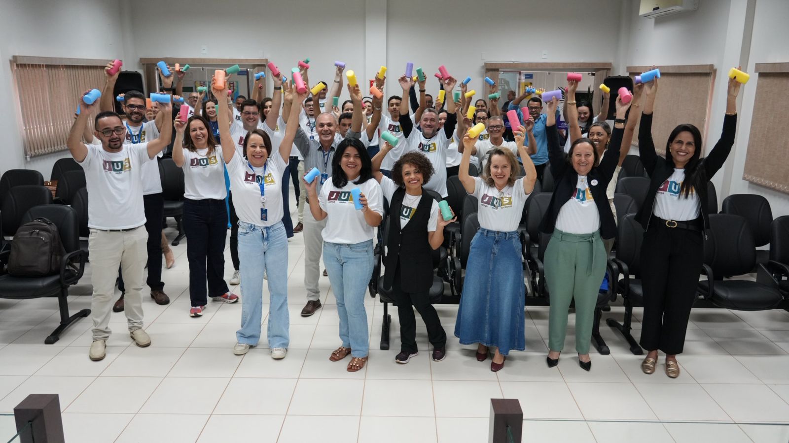 A imagem mostra um grupo de pessoas sorridentes, vestindo camisetas do projeto “JUS em Ação”, levantando objetos coloridos durante uma dinâmica realizada com servidores da Comarca de Novo Acordo. 