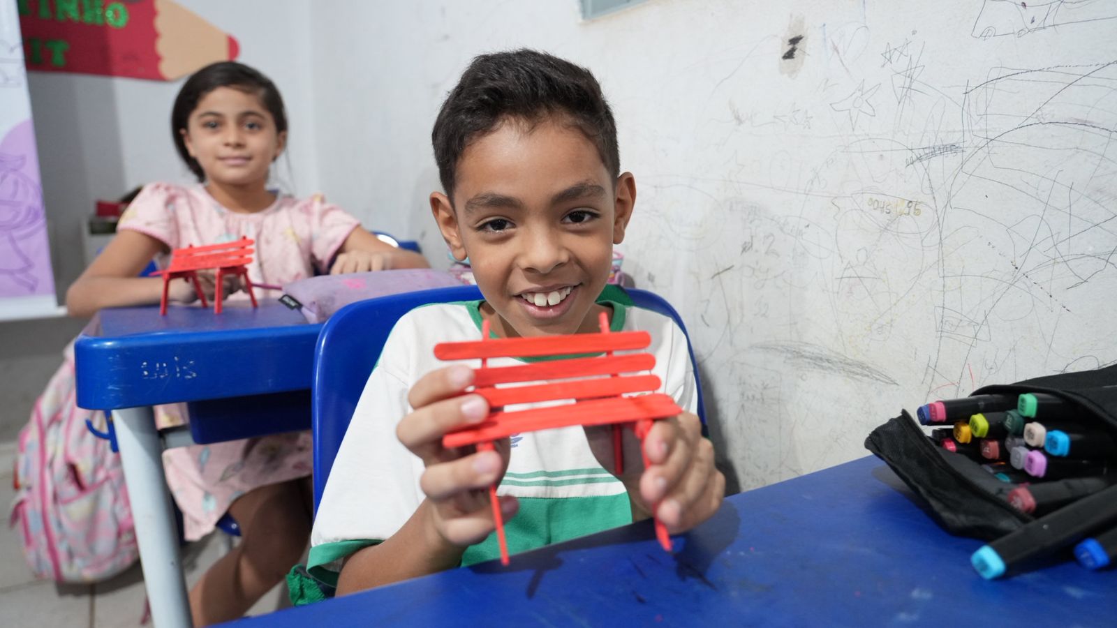 Menino sorrir pra foto segurando um banquinho vermelho; ele está sentado em carteira azul; ao fundo uma menina