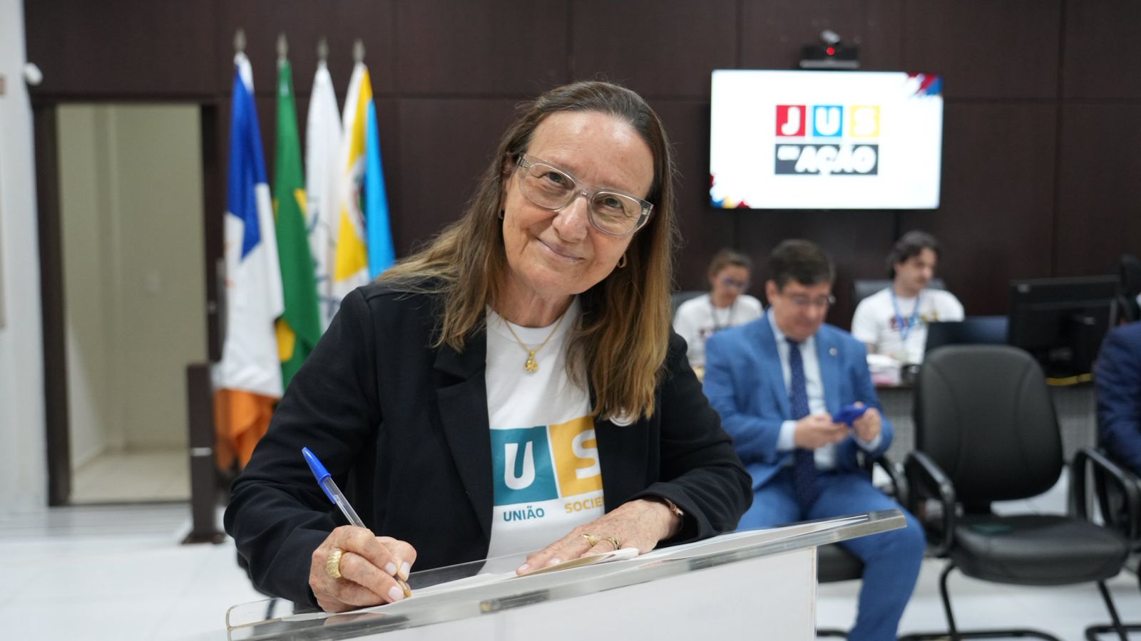 Presidente do TJTO, uma mulher de pele branca, cabelos claros, de óculos, terno preto e camisa branca com o nome JUS, olha pra foto ao assinar documento