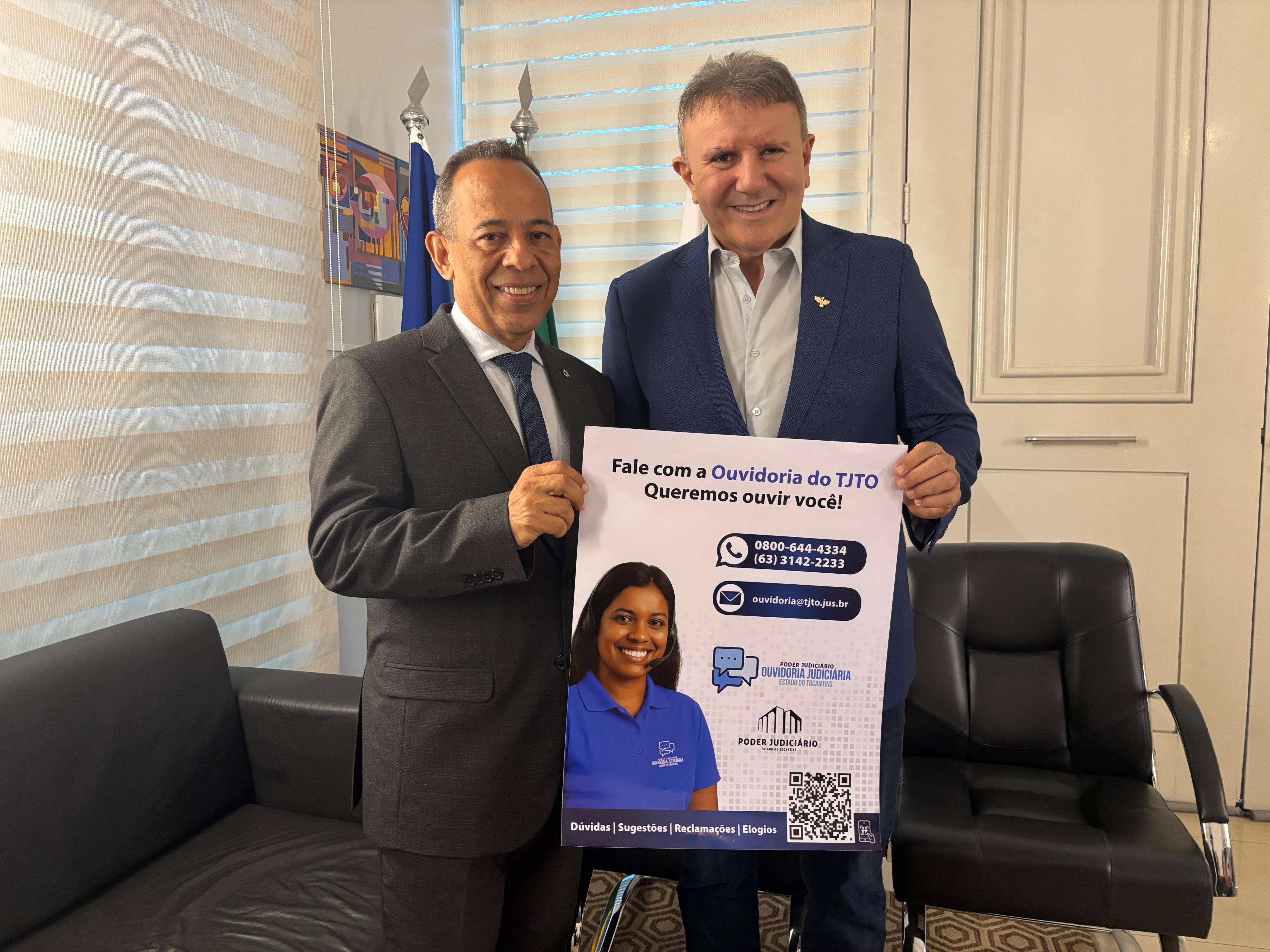 A imagem mostra o ouvidor judiciário, desembargador João Rodrigues, e o prefeito de Palmas, Eduardo Siqueira Campos, sorrindo e posando juntos enquanto seguram um cartaz publicitário da Ouvidoria do Tribunal de Justiça do Tocantins (TJTO), com os canais de atendimento destacados.