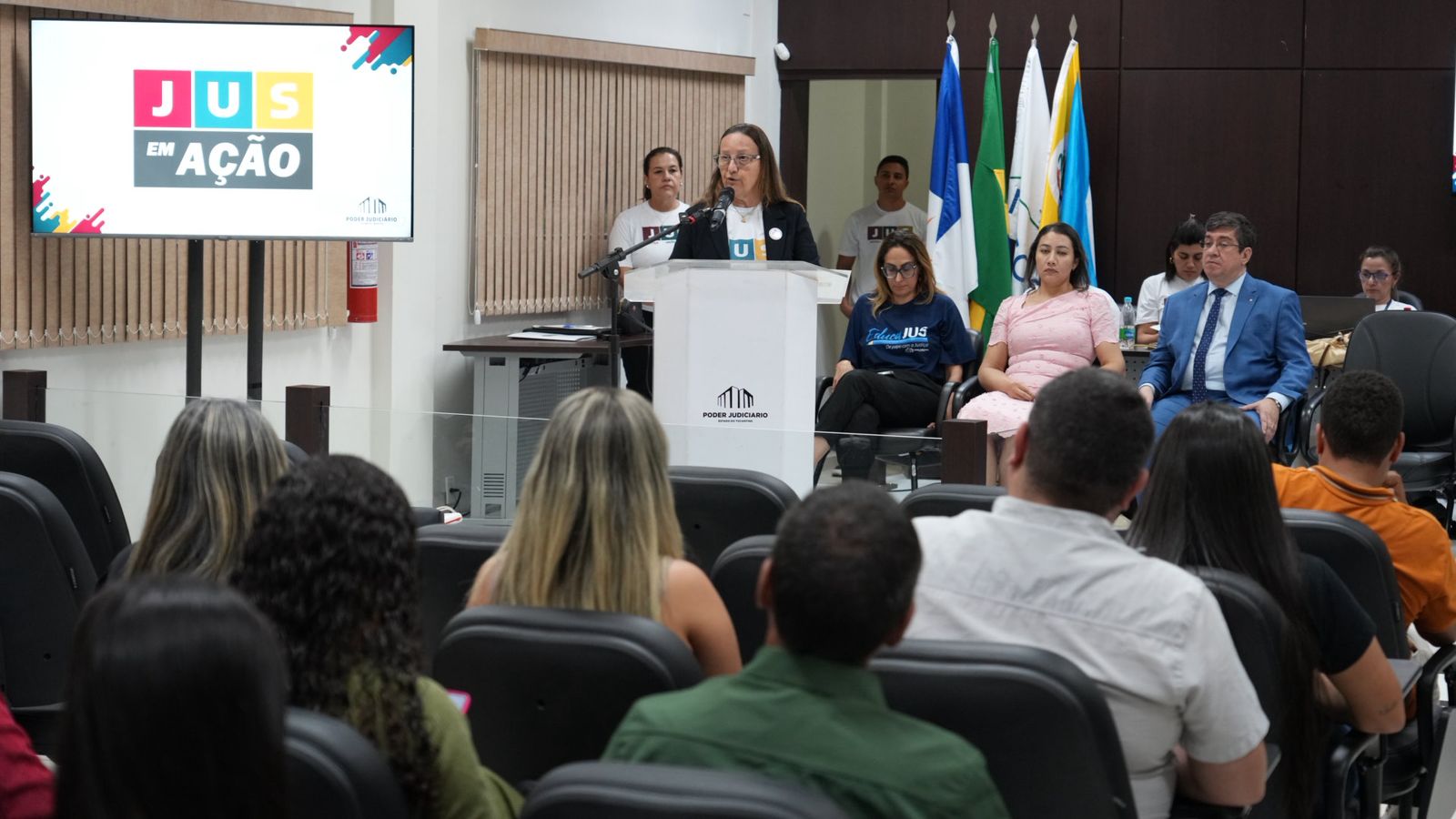 A imagem mostra a presidente do Tribunal de Justiça do Tocantins, desembargadora Maysa Vendramini Rosal, discursando ao microfone em um púlpito durante o projeto JUS em Ação. À sua frente, várias pessoas acompanham atentamente, enquanto à esquerda estão autoridades sentadas e, ao fundo, quatro bandeiras compõem o cenário. No lado direito, uma TV exibe a logomarca do projeto.