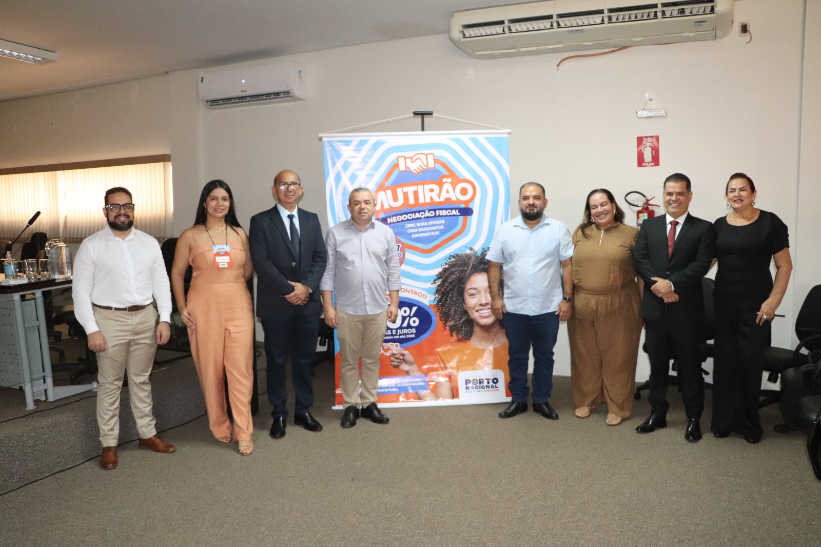A foto mostra um grupo de nove pessoas posando lado a lado em um ambiente interno, como uma sala de reuniões, no Fórum de Porto Nacional. Ao centro, atrás do grupo, há um banner colorido com o texto “Mutirão – Negociação Fiscal”