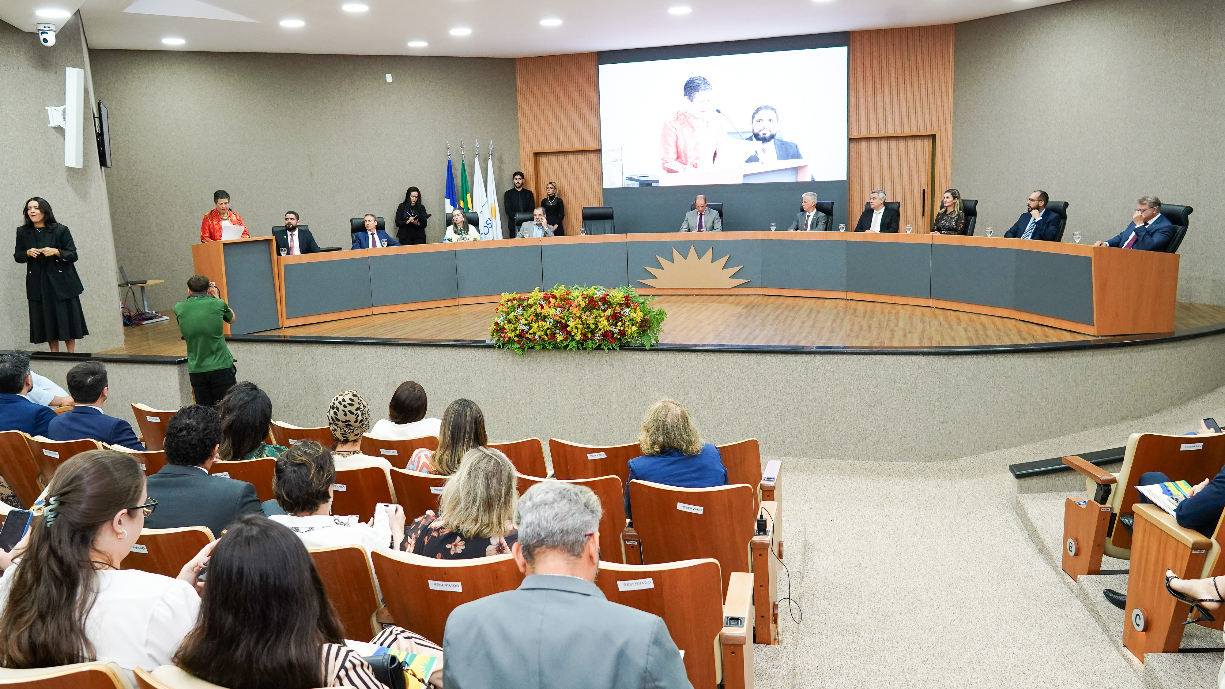 Na imagem, vê-se um auditório com público sentado enquanto uma cerimônia oficial acontece no palco. Uma mesa de honra com diversas autoridades está disposta ao fundo, e uma oradora fala ao púlpito, com uma intérprete de Libras posicionada à esquerda. Um fotógrafo registra o momento, e um telão ao fundo projeta a imagem da fala.