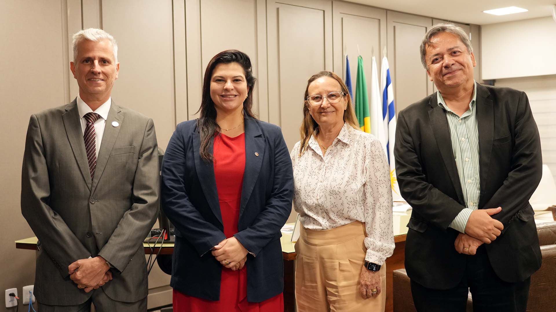 Na imagem, quatro pessoas posam para foto em ambiente institucional. Elas estão alinhadas lado a lado, vestindo roupas formais, com bandeiras ao fundo.