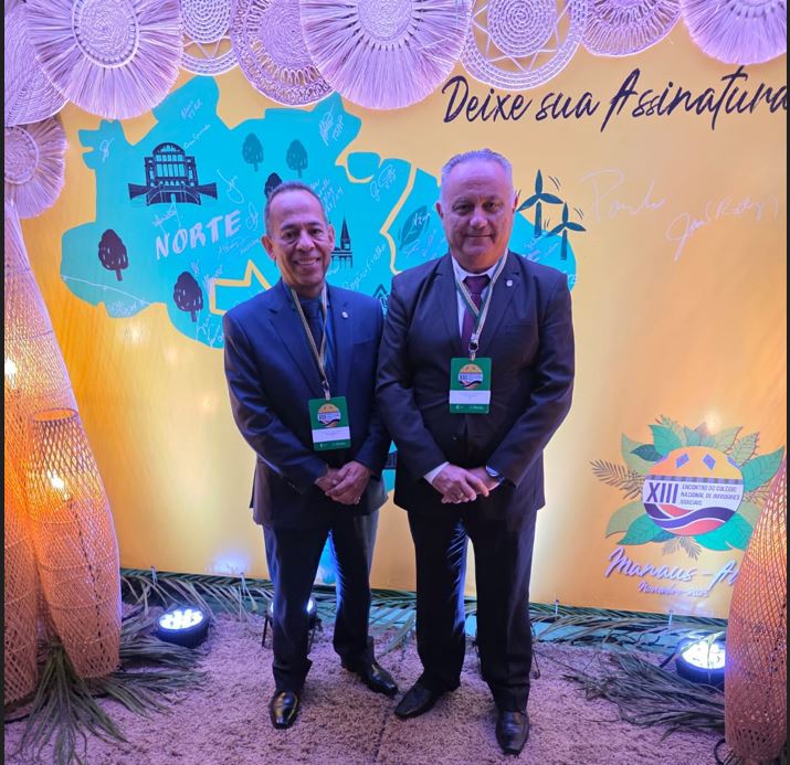 Dois homens, ambos de terno e crachá de evento, posam lado a lado diante de um painel iluminado com o mapa da região Norte, elementos gráficos e a frase “Deixe sua Assinatura”. No canto inferior direito há o logo do XIII Encontro do Colégio Nacional de Ouvidores Judiciais.