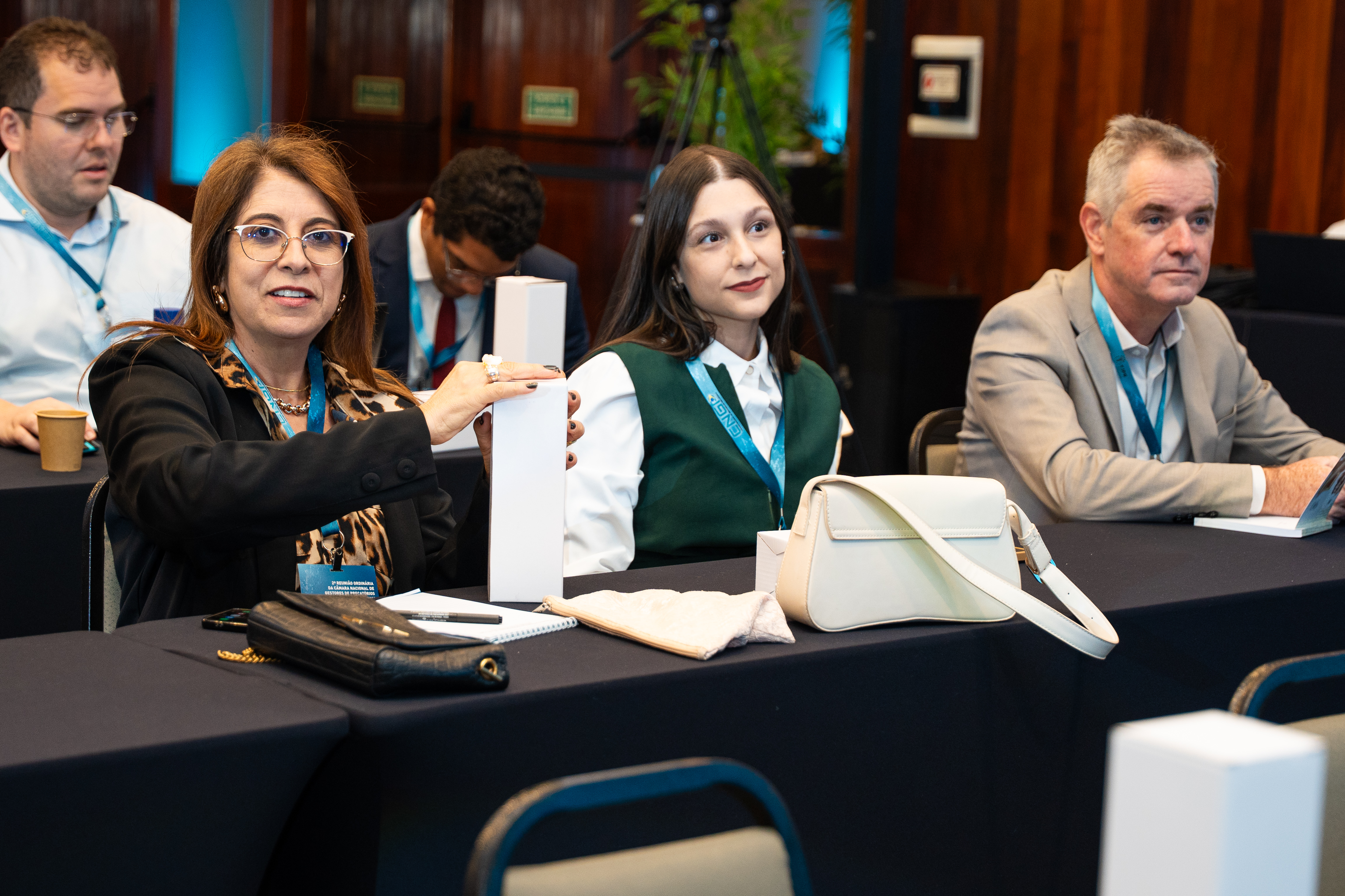 Em uma sala de conferências, três pessoas estão sentadas lado a lado. À esquerda, uma mulher de óculos e blazer preto segura uma caixa branca. Ao centro, outra mulher de colete verde e camisa branca observa atenta. À direita, um homem de terno bege acompanha a atividade. Sobre a mesa, há bolsas e cadernos. O ambiente é formal e iluminado.