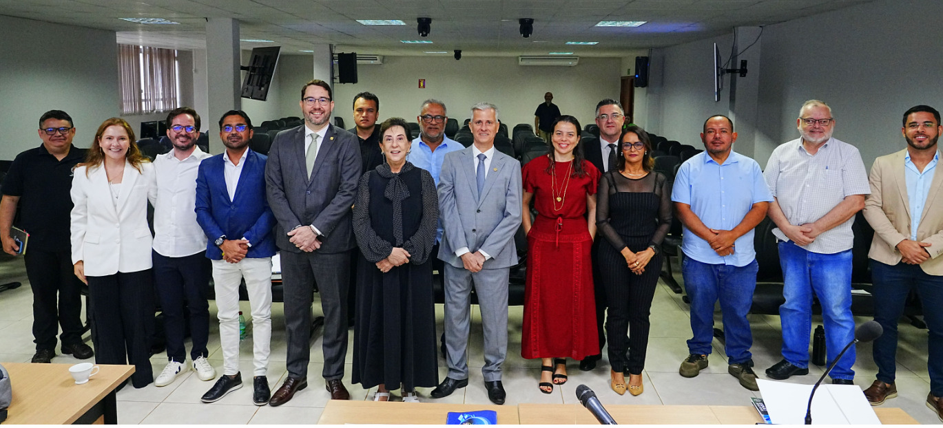 Um grande grupo de participantes posa para foto oficial em um auditório, de pé em frente a um painel digital do evento.