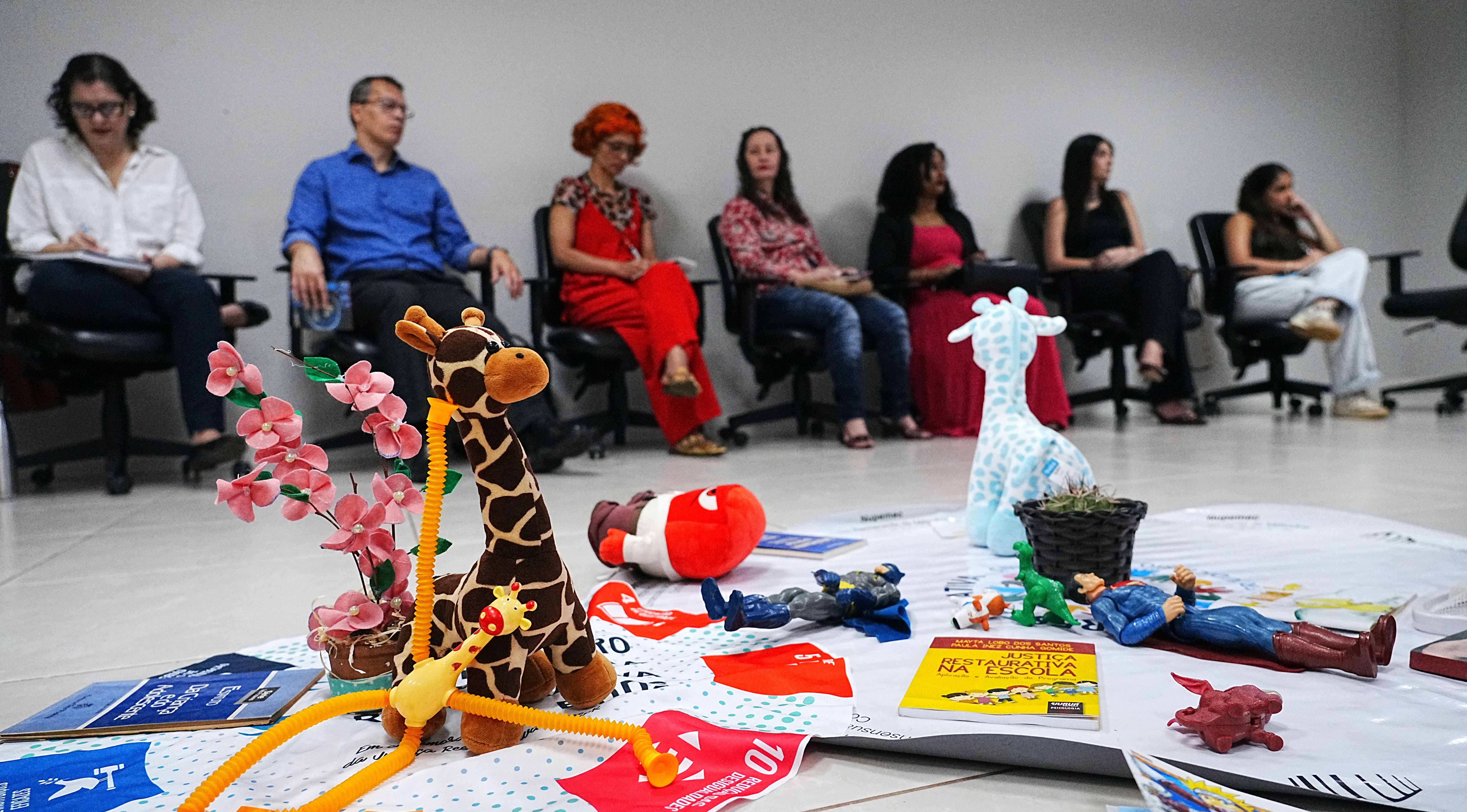Um grupo de pessoas está sentado em círculo, observando uma dinâmica ao centro do espaço. No chão, há brinquedos, livros e objetos simbólicos utilizados na atividade.