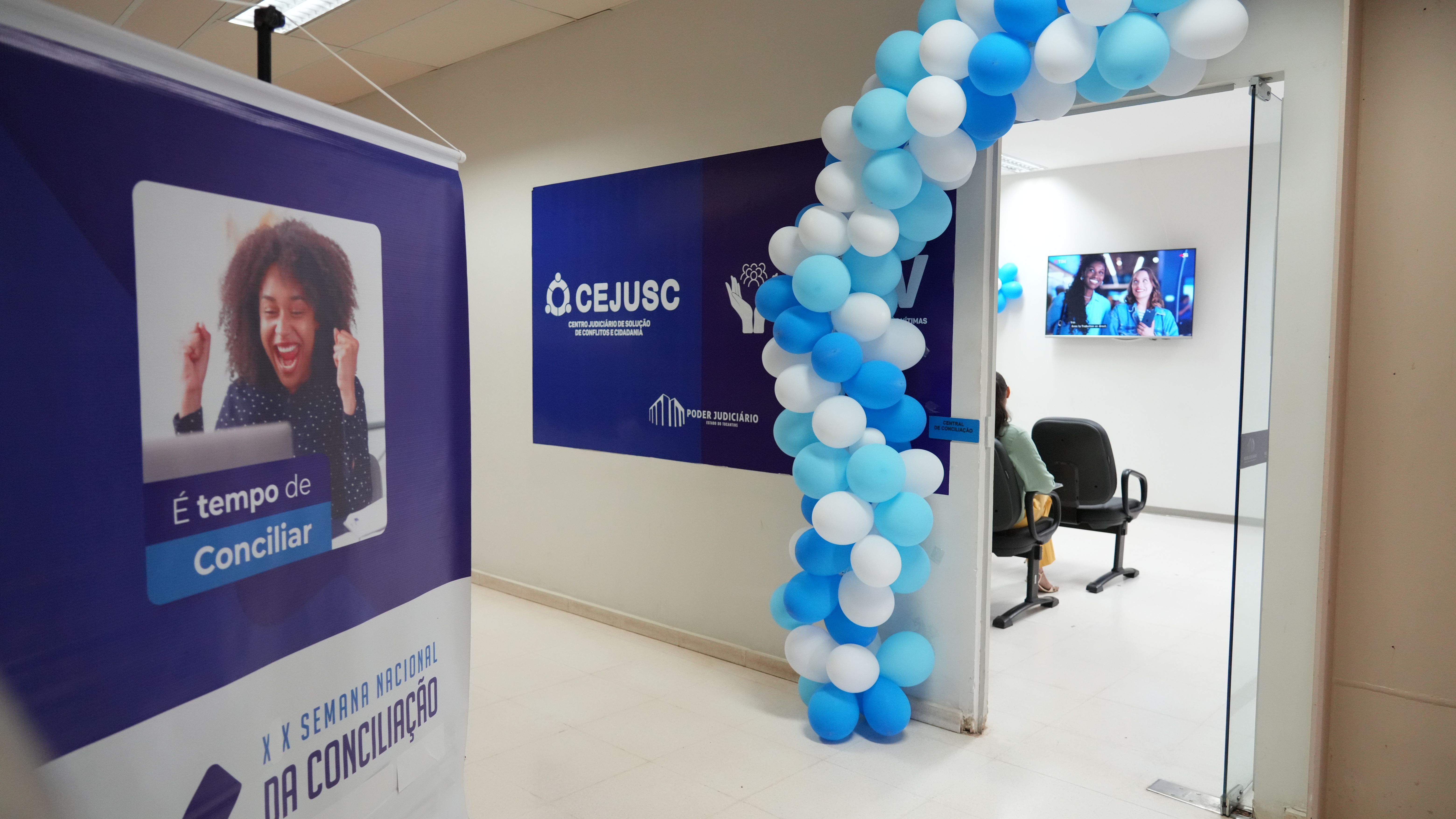 Uma entrada decorada com balões em tons de azul e branco dá acesso a uma sala do Cejusc. À esquerda, há um banner da Semana Nacional da Conciliação com a mensagem “É tempo de conciliar”. Dentro da sala, cadeiras e uma televisão com vídeo em exibição completam o ambiente.