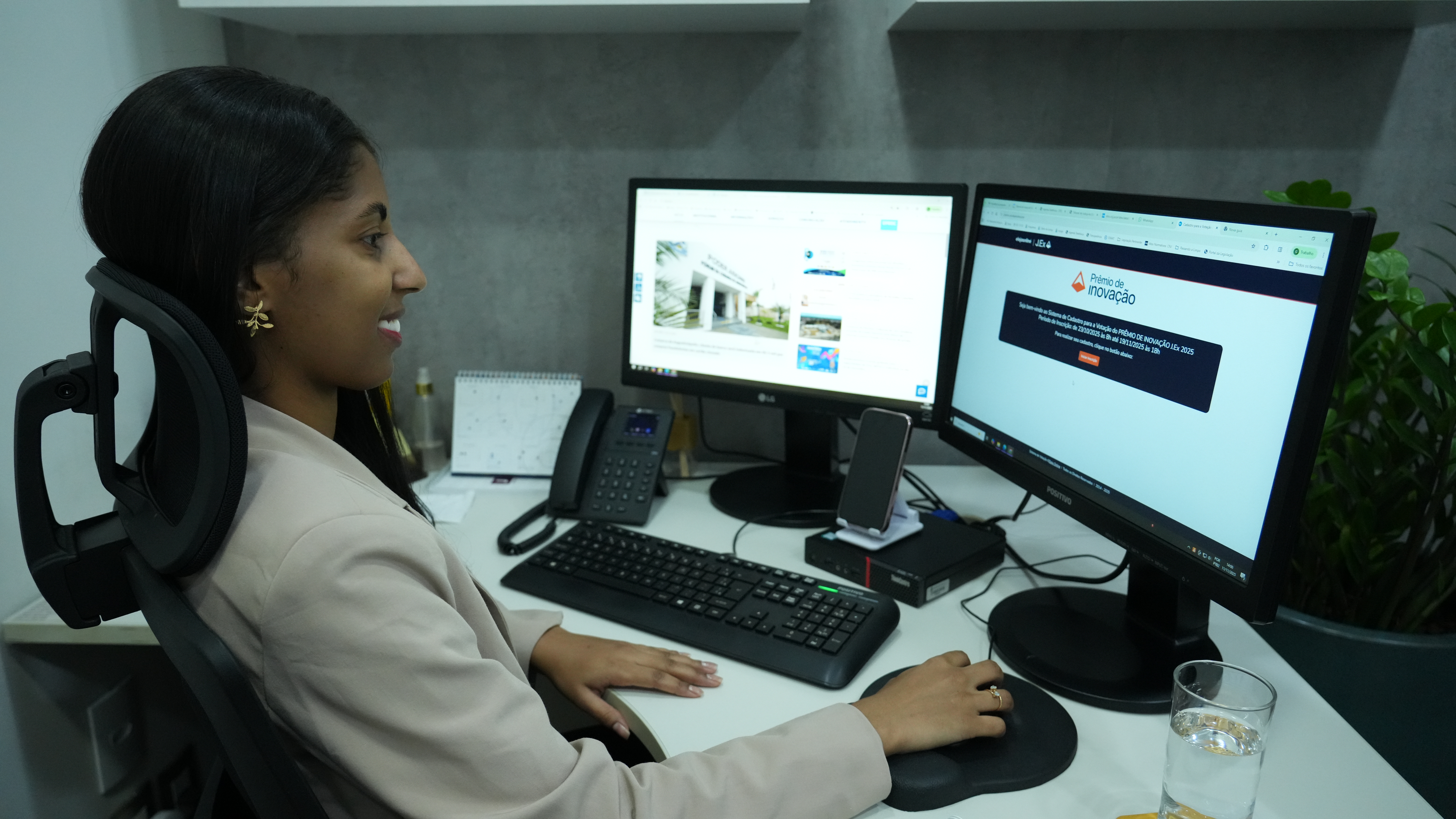 Uma mulher jovem está sentada em uma cadeira de escritório, usando computador. Ela sorri enquanto trabalha em duas telas: uma mostra um site institucional e a outra exibe a página de votação do “Prêmio de Inovação”. Na mesa, há um teclado, telefone fixo, celular apoiado em um suporte e um copo com água. Ao fundo, há uma parede cinza e uma planta. A cena mostra um ambiente de trabalho organizado e moderno.
