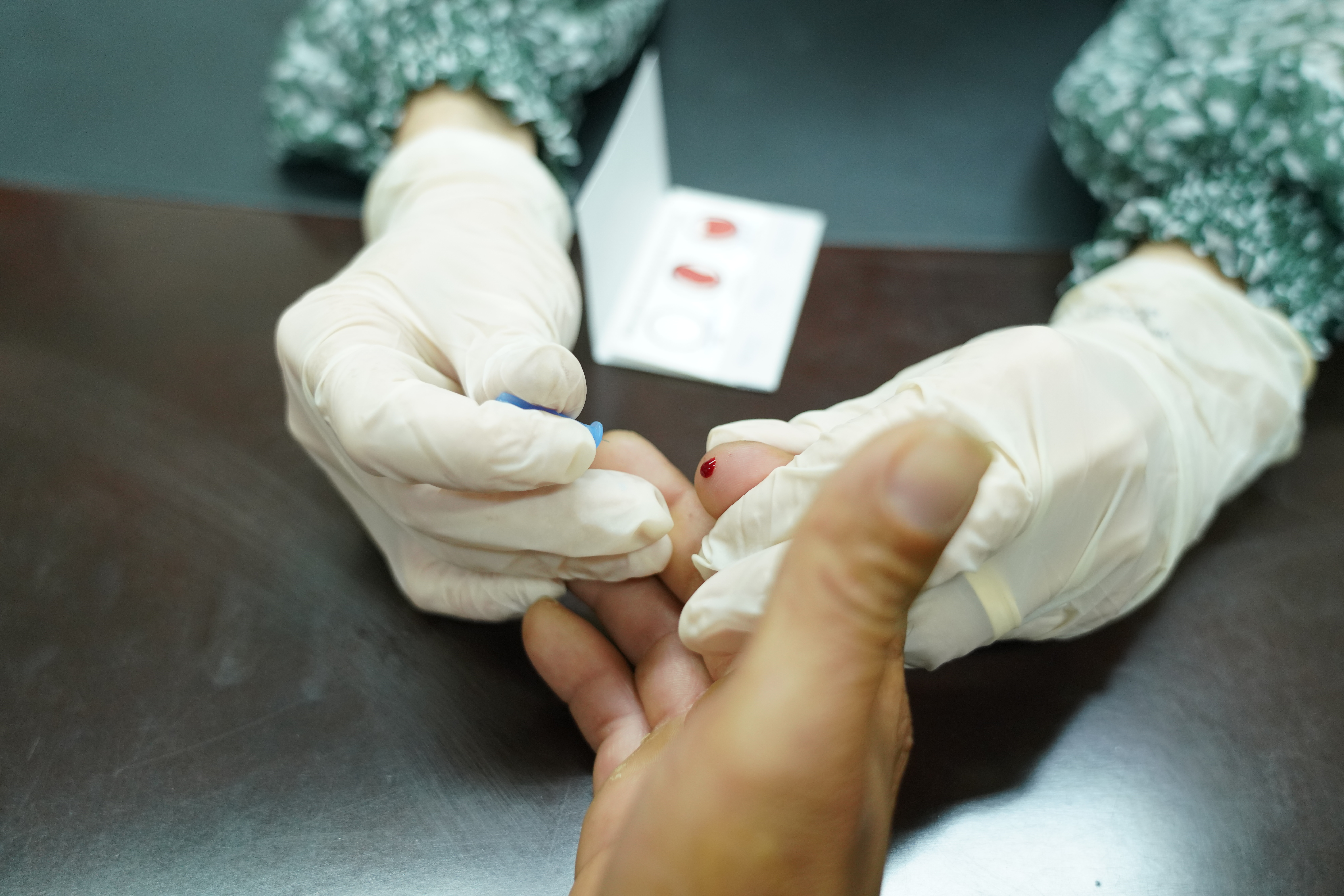 A imagem mostra um(a) profissional da saúde usando luvas descartáveis enquanto realiza a coleta de uma pequena amostra de sangue da ponta do dedo de uma pessoa, utilizando uma lanceta. Uma gota de sangue é visível no dedo do paciente, e ao fundo, sobre a mesa, há um cartão com espaços marcados para o teste, possivelmente um exame rápido. A cena se passa sobre uma superfície escura, com foco nas mãos e na coleta do sangue.