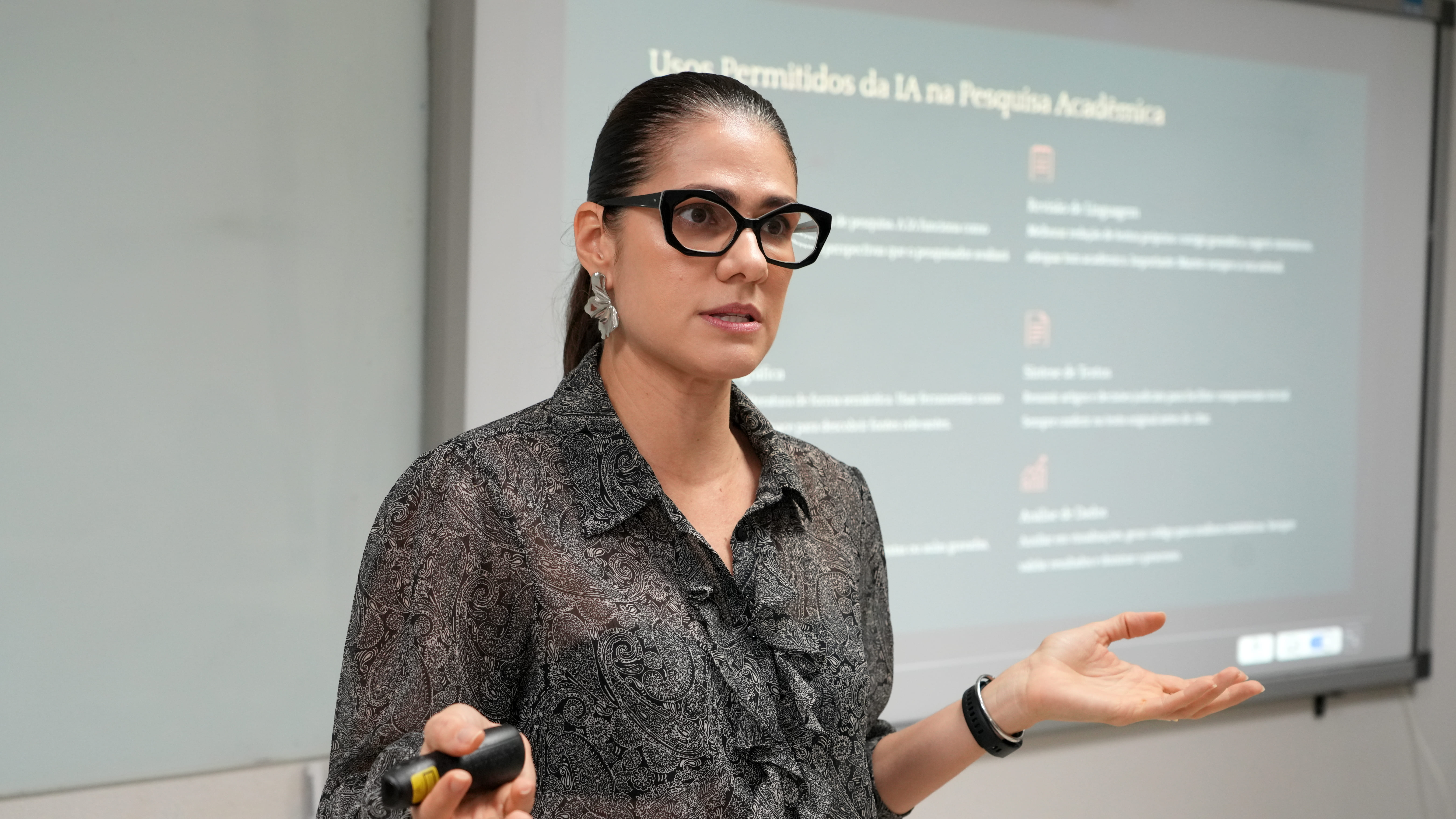 Mulher fala durante apresentação em sala de aula, com projeção de slide ao fundo sobre o uso da inteligência artificial na pesquisa acadêmica.