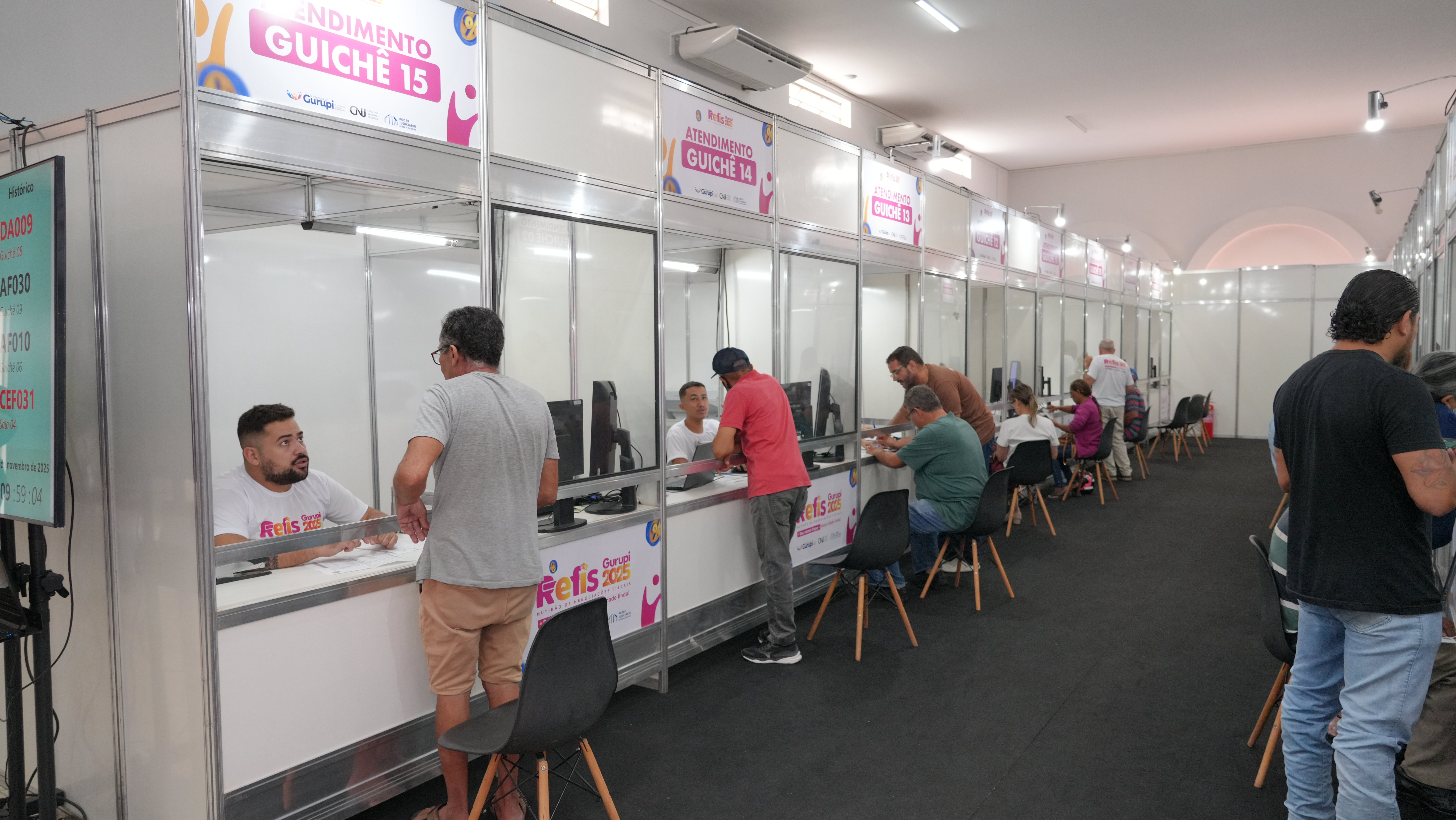 Imagem mostra o espaço de atendimento do Mutirão de Negociação Fiscal de Gurupi (Refis 2025), com vários guichês em funcionamento. Servidores e contribuintes conversam e formalizam acordos para regularização de débitos municipais em um ambiente organizado e sinalizado.
