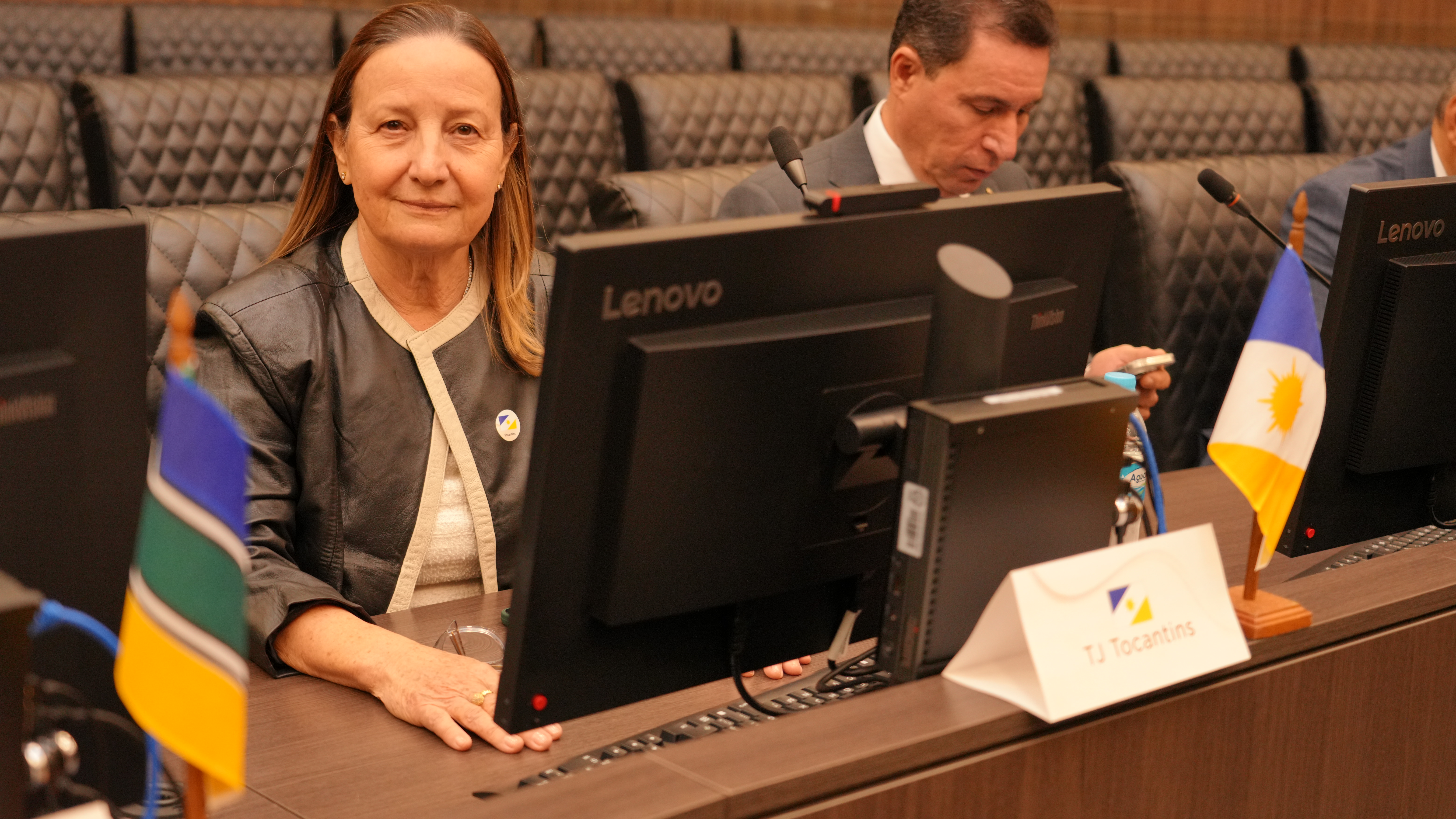 Presidente do TJTO, desembargadora Maysa está sentada em uma mesa de reunião institucional, diante de um computador, olhando para a câmera. Ao lado, outro participante manuseia o celular. Sobre a mesa, há microfones, placas de identificação do TJTO e pequenas bandeiras.