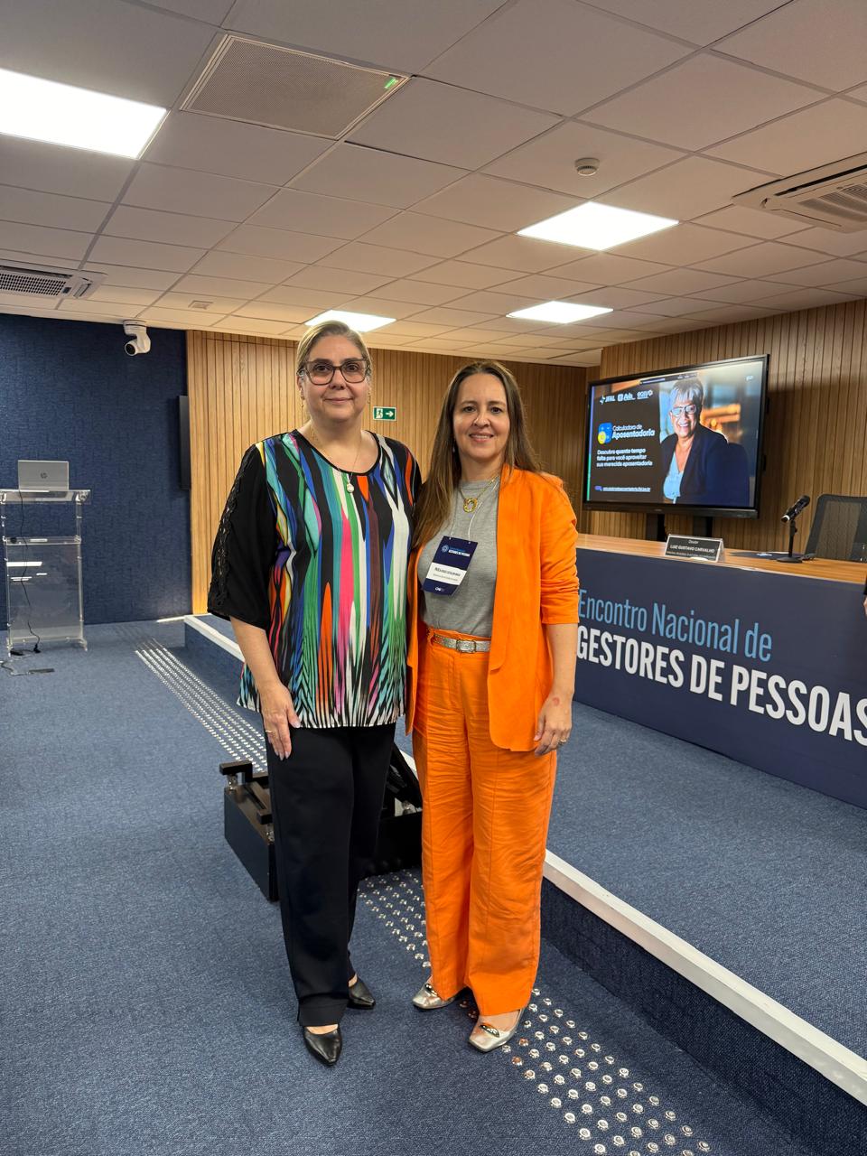 Juíza Flávia Bovo e Paula Maia posam para a foto em um auditório. À esquerda, a juíza usa blusa colorida de mangas pretas e calça escura; à direita, Paula Maia veste um conjunto laranja com camiseta clara e crachá. Ao fundo, há um palco com mesa, microfone, telão ligado e o painel do evento “Encontro Nacional de Gestores de Pessoas”.