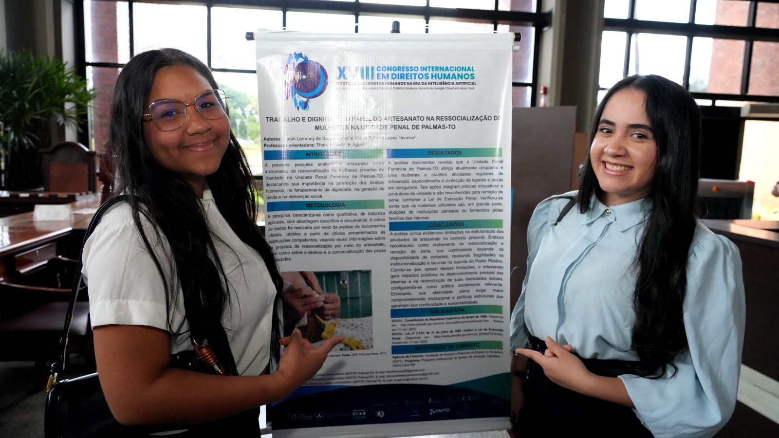 A foto mostra duas jovens posando ao lado de um pôster científico exposto no XVIII Congresso Internacional em Direitos Humanos. Elas sorriem e apontam para o painel, que apresenta uma pesquisa sobre o papel do artesanato na ressocialização de mulheres na Unidade Penal Feminina de Palmas.
