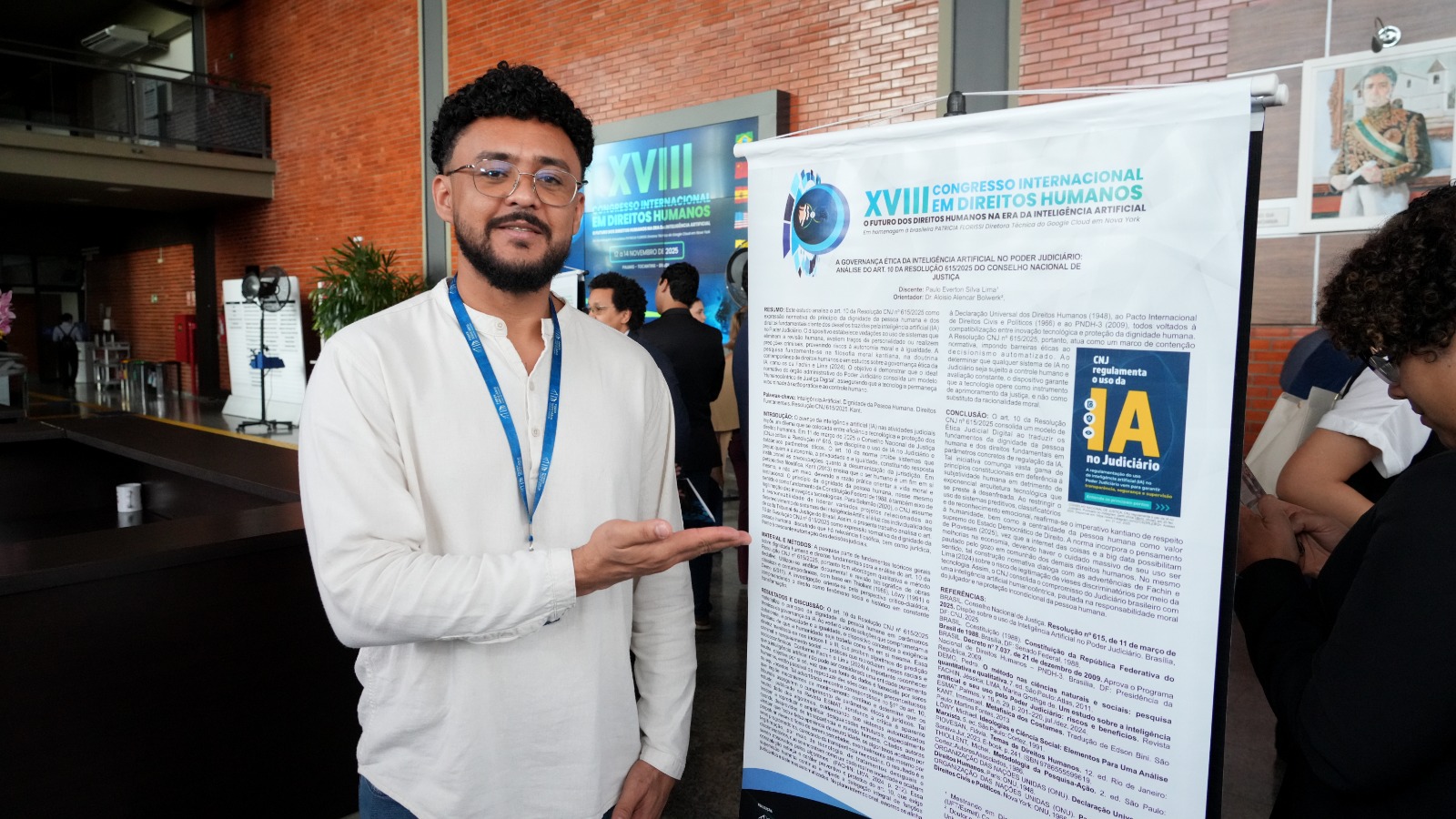 Na imagem, estudante usa camisa clara e crachá azul do Poder Judiciário enquanto aponta para um pôster científico exposto em painel vertical. O pôster apresenta texto extenso sobre direitos humanos e inteligência artificial, com o logo do XVIII Congresso Internacional em Direitos Humanos ao topo. Ao fundo, há pessoas circulando pelo hall e uma parede com revestimento cerâmico.parentes.