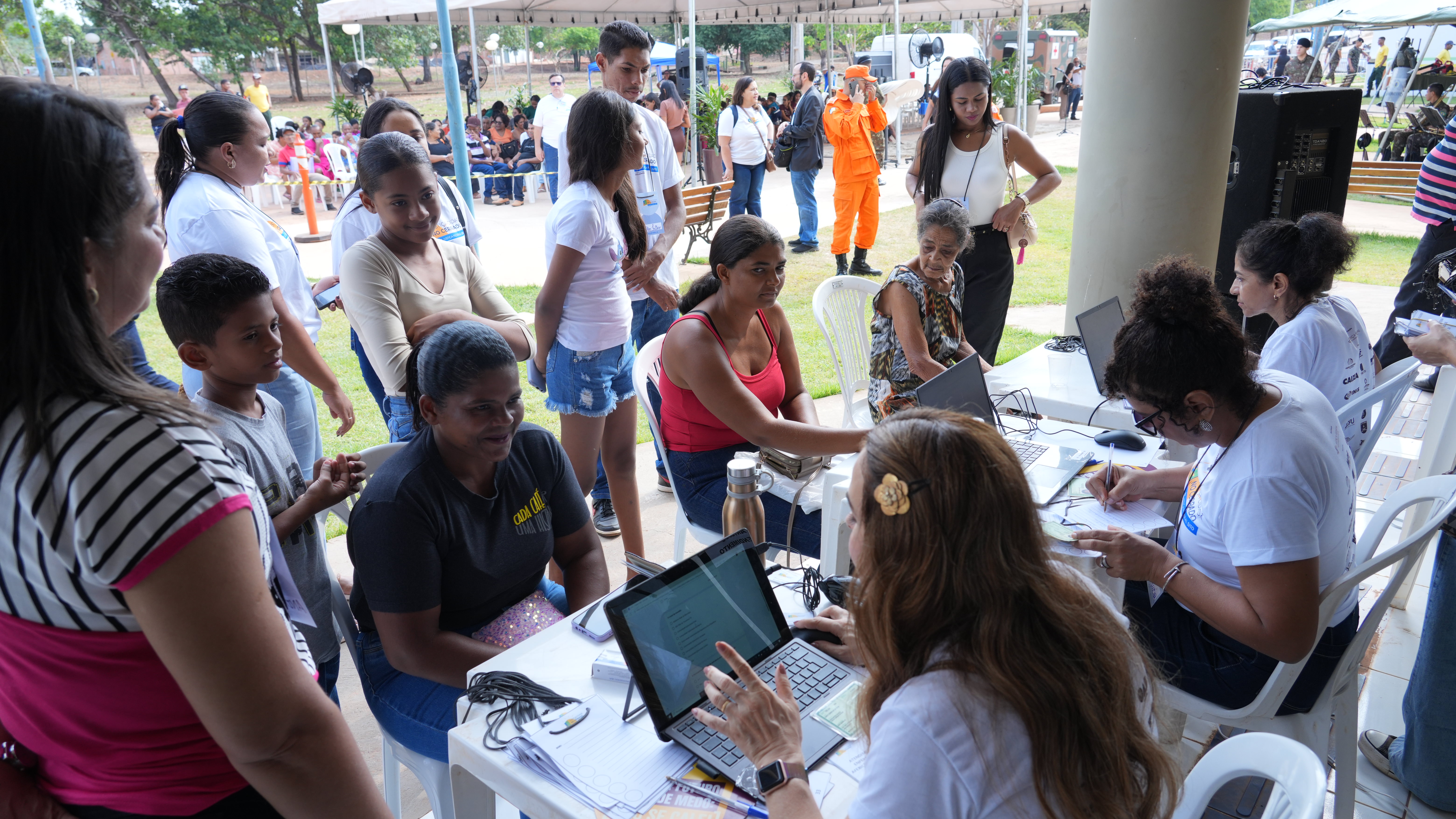 A imagem mostra um atendimento comunitário ao ar livre, com várias pessoas sentadas e em fila, enquanto servidoras realizam cadastros em laptops. Em primeiro plano, mulheres e crianças aguardam e conversam. No fundo, há uma área coberta com mais pessoas participando da ação.