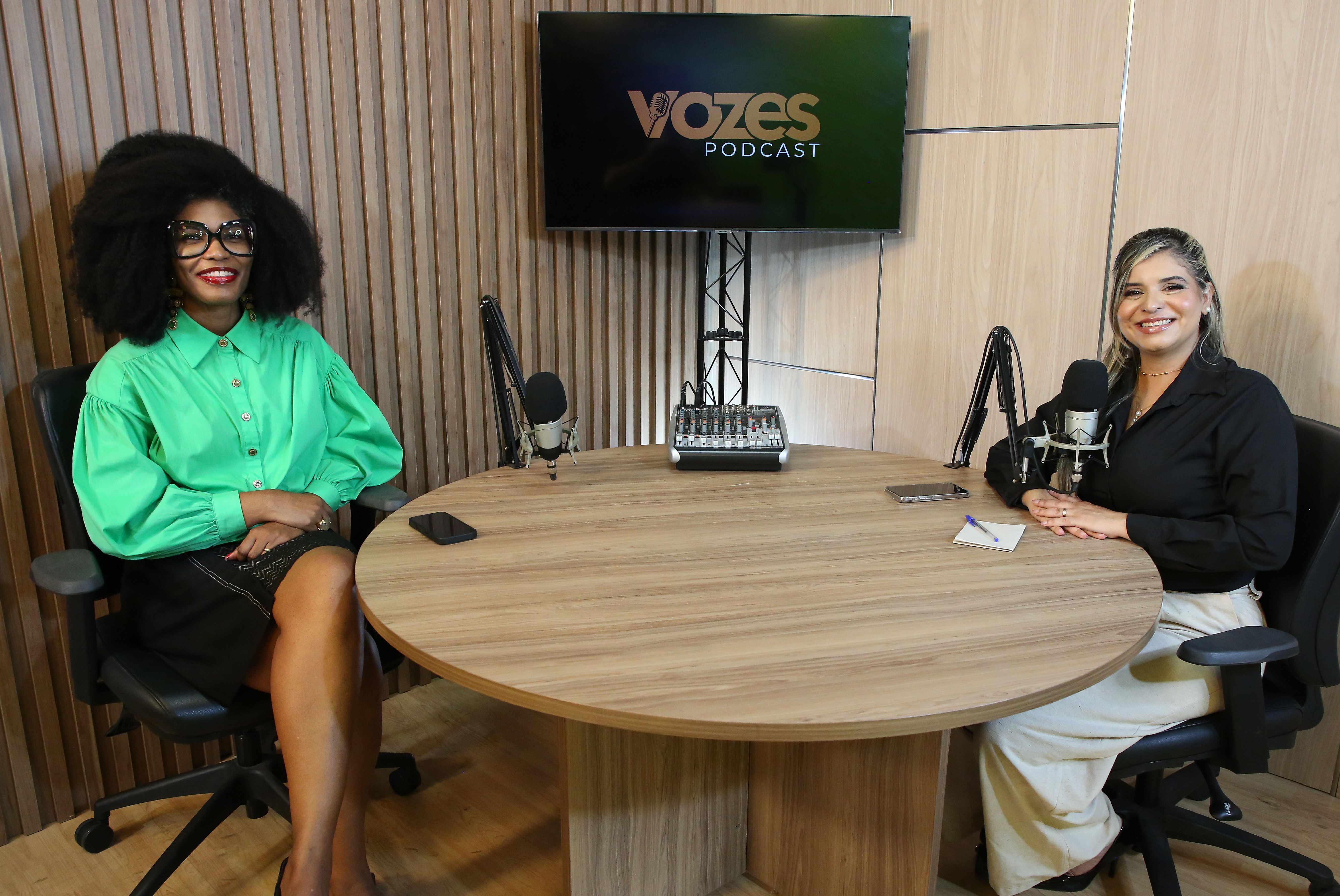 A foto mostra duas mulheres sentadas em um estúdio de podcast, posicionadas em lados opostos de uma mesa redonda de madeira. À esquerda, está uma mulher de pele retinta, com cabelo afro volumoso, óculos grandes de armação preta e camisa verde vibrante. À direita, uma mulher de pele clara e cabelo loiro preso parcialmente, usando blusa preta e calça clara, segura um microfone enquanto sorri. Ao fundo, uma TV exibe o nome “Vozes Podcast”, compondo o cenário profissional da gravação.