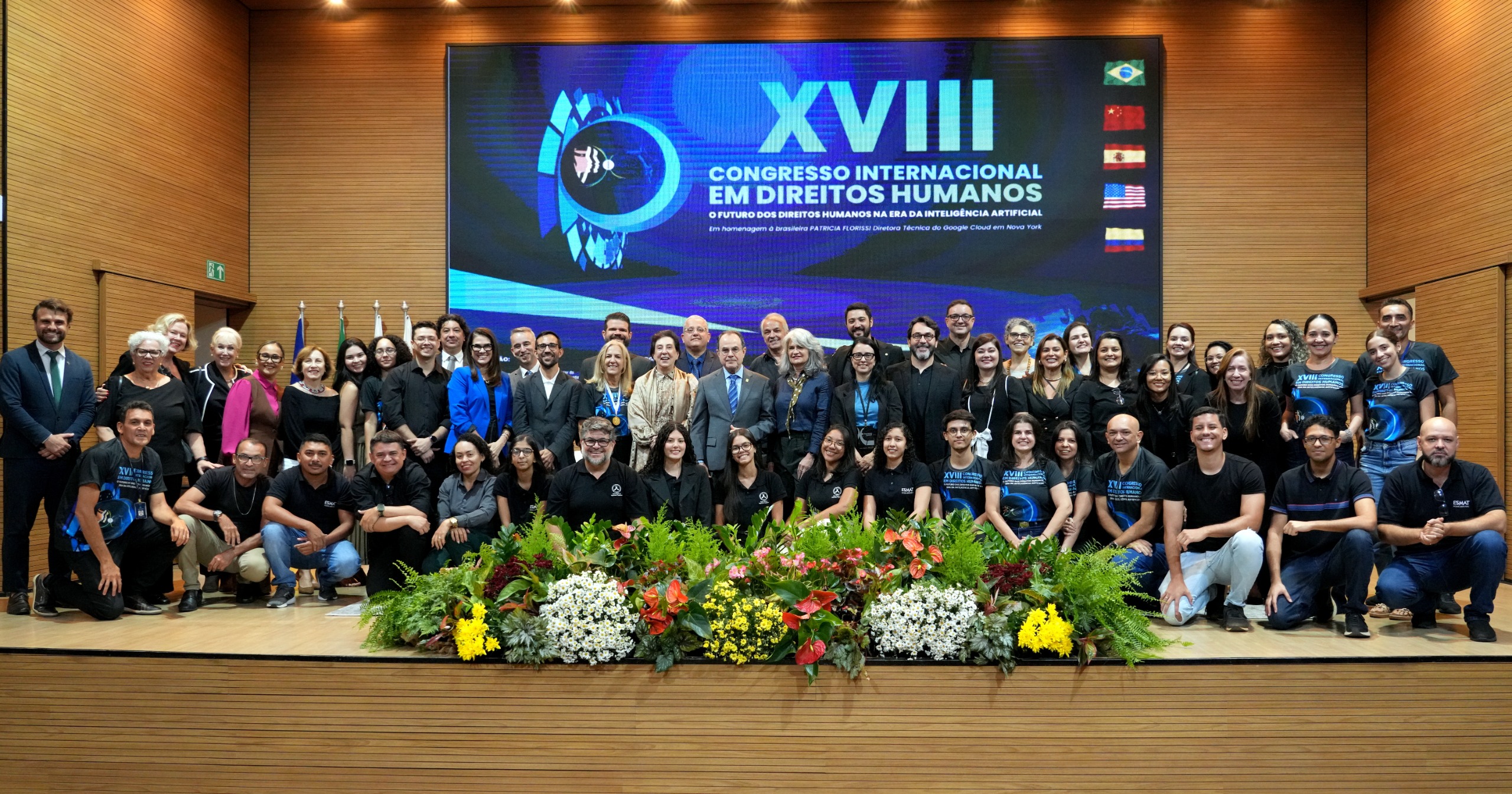 Foto oficial do XVIII Congresso Internacional em Direitos Humanos, mostrando um grande grupo de participantes reunidos no palco do auditório. Ao fundo, um painel eletrônico exibe o nome do evento. À frente do grupo há um arranjo de flores com cores fortes, como amarelo, vermelho e branco. O ambiente é amplo, com paredes de madeira e iluminação clara, reforçando o caráter institucional do encontro.