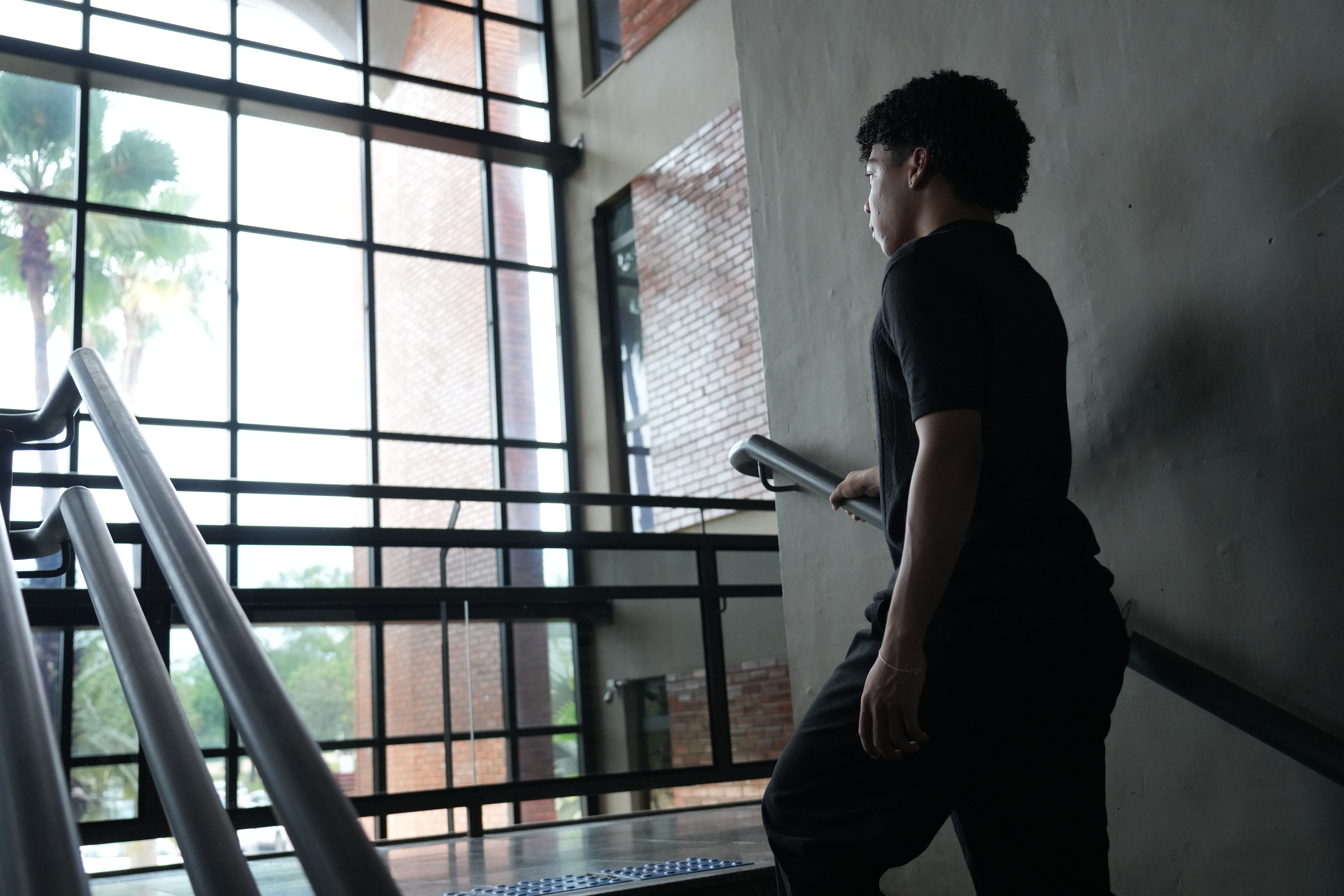 A foto mostra um jovem subindo uma escada interna, segurando o corrimão. O espaço tem parede cinza e uma grande fachada de vidro ao fundo, por onde entram luz natural e a vista de árvores e prédios de tijolos.