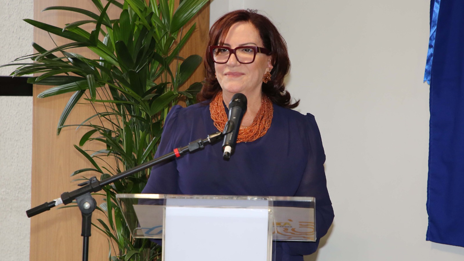 A foto mostra a desembargadora Etelvina falando ao microfone em um púlpito transparente. Ela usa óculos, colar volumoso e blusa azul. Ao fundo, há uma planta decorativa e parte de uma cortina azul.