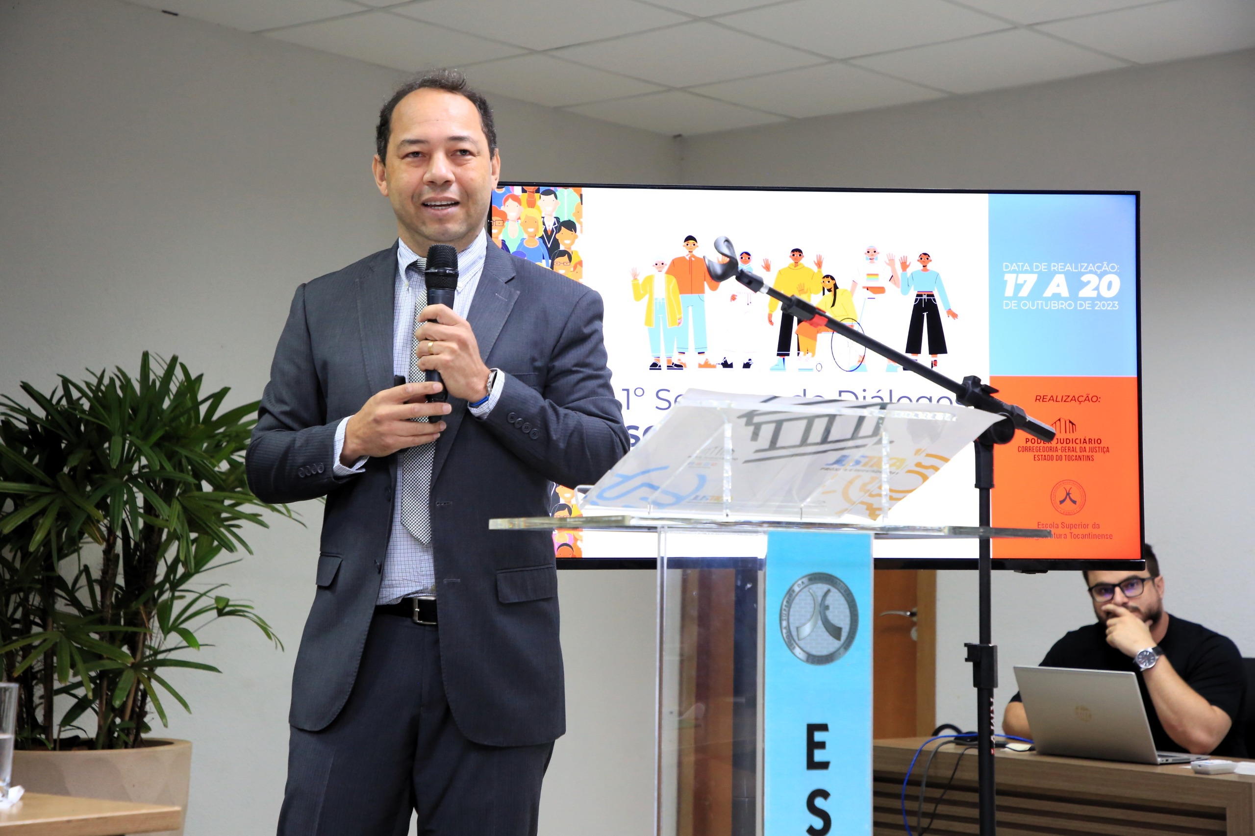 A foto mostra o juiz Jean, de terno, falando ao microfone em um auditório. À frente dele há um púlpito transparente com identificação institucional. Atrás, uma tela exibe informações sobre um evento. À direita, um homem está sentado diante de um notebook.